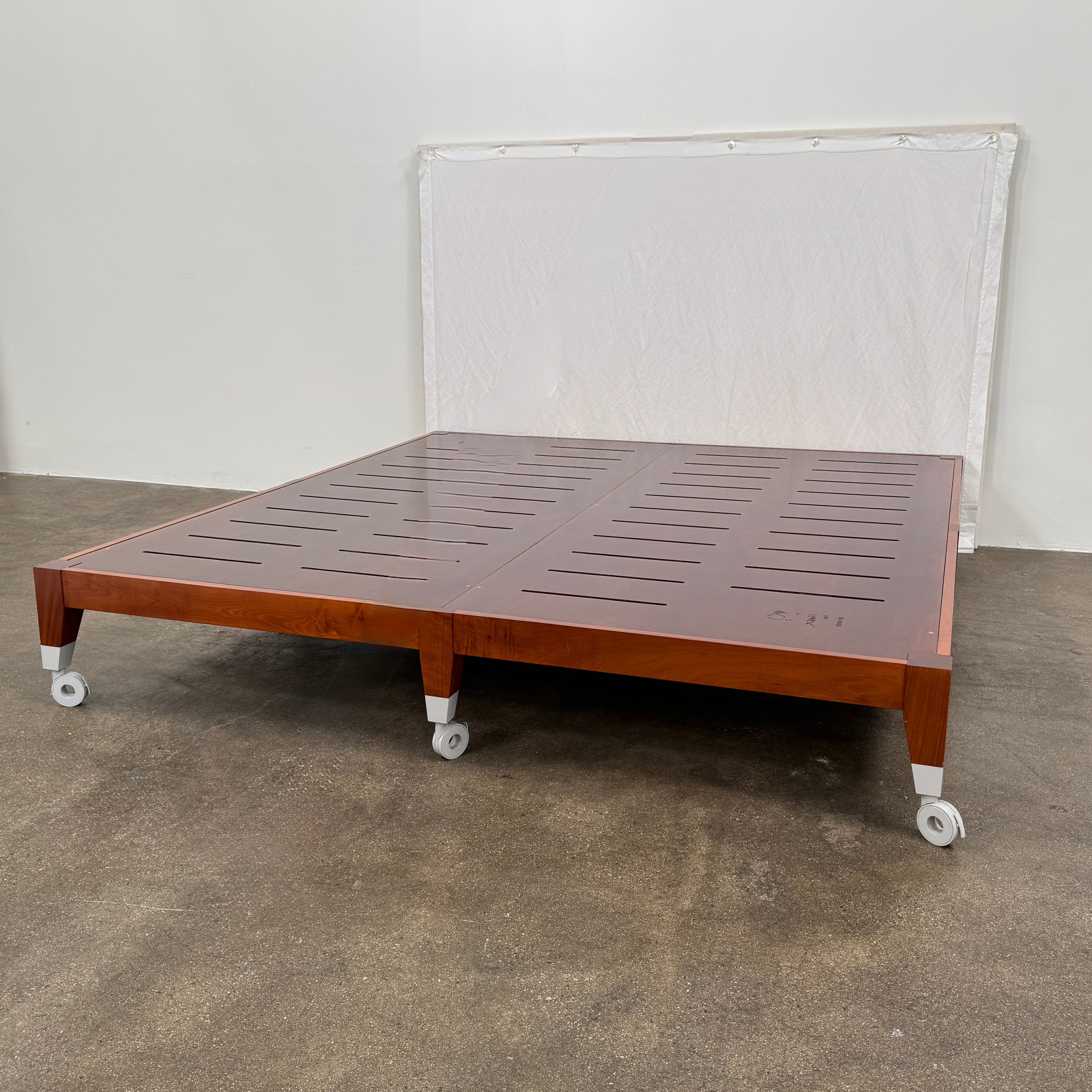 The Driade Neoz European King Size Bed by Driade, featuring wooden slats, wheels, and a white fabric headboard, stands on a concrete floor against a plain white wall.
