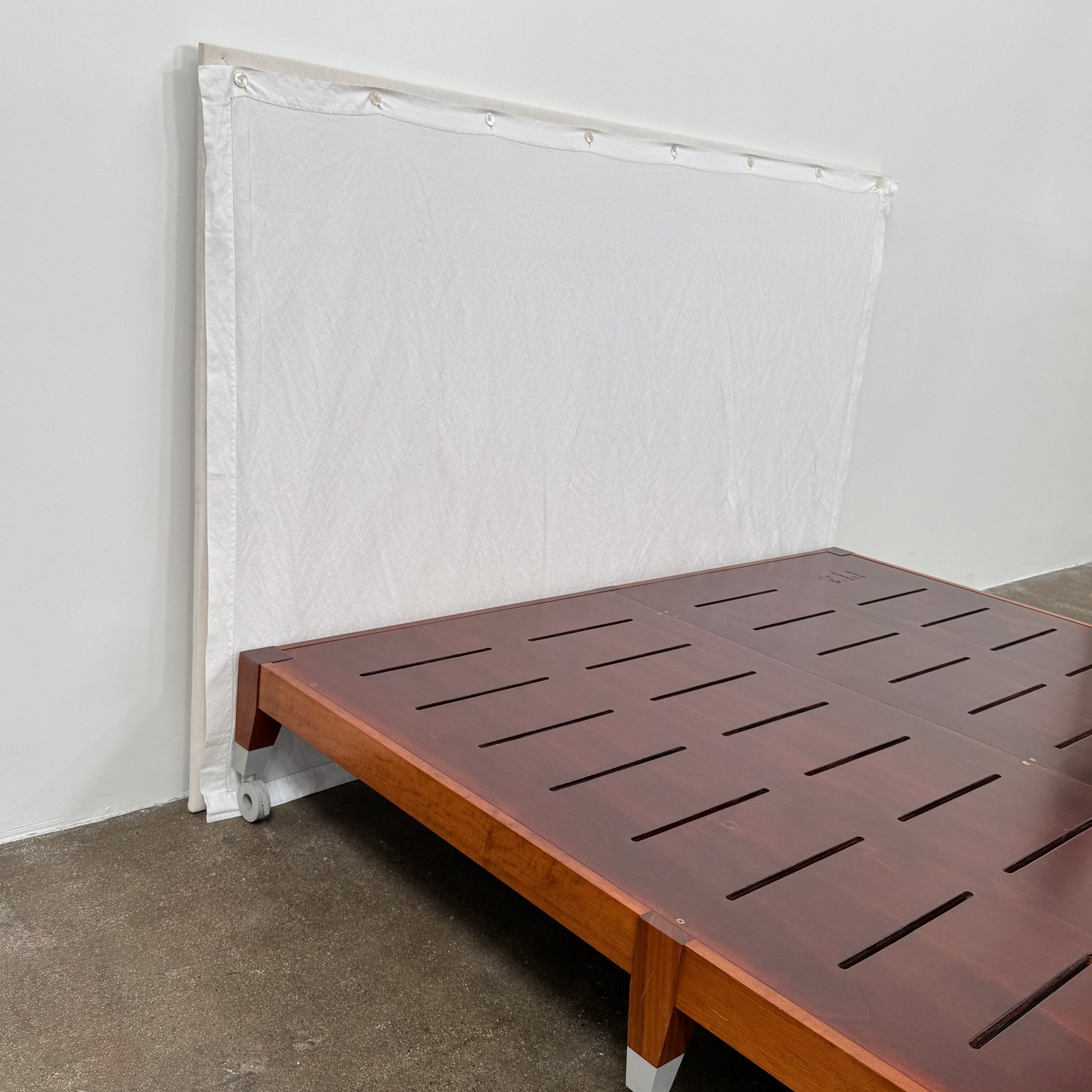 The Driade Neoz European King Size Bed by Driade, featuring wooden slats, wheels, and a white fabric headboard, stands on a concrete floor against a plain white wall.
