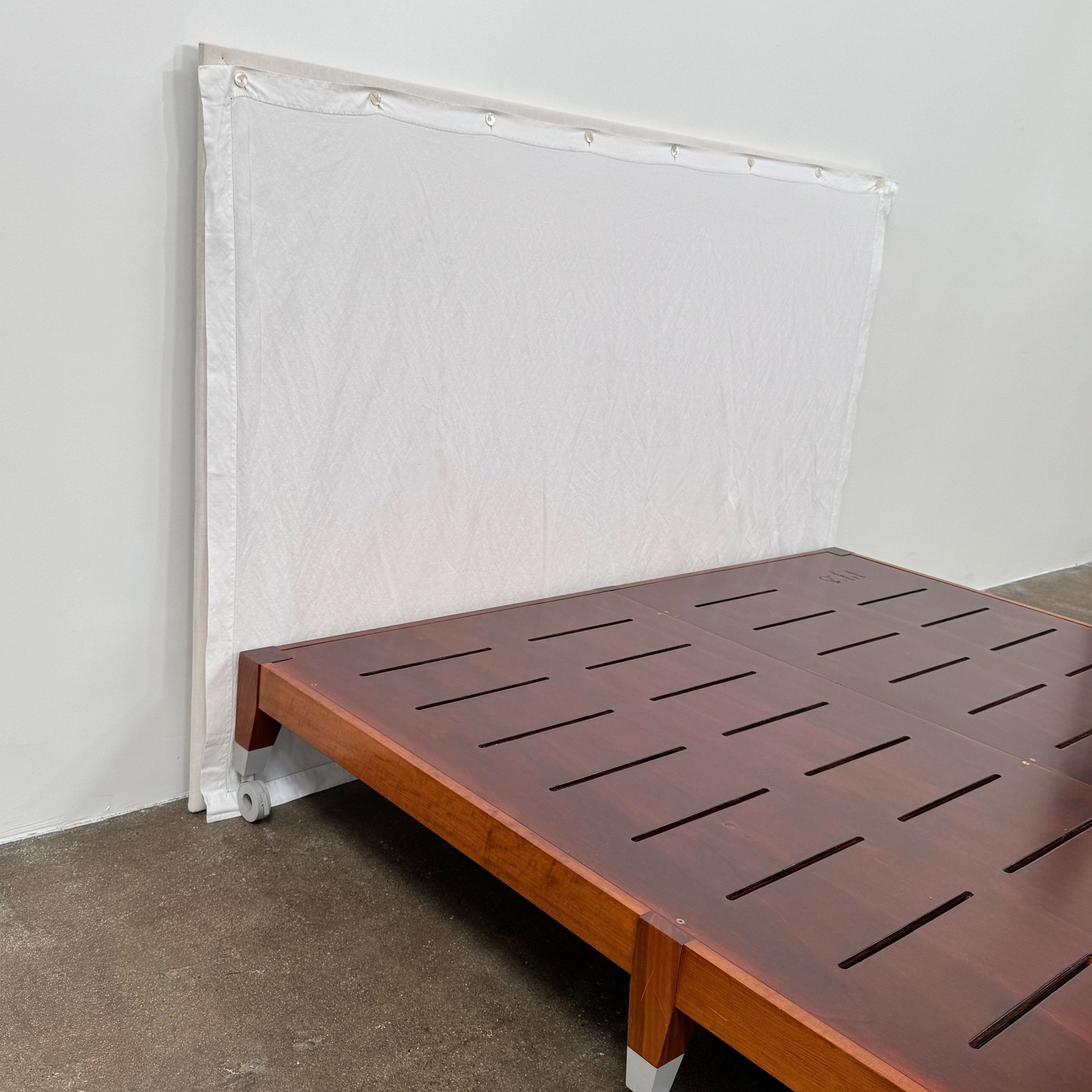 The Driade Neoz European King Size Bed by Driade, featuring wooden slats, wheels, and a white fabric headboard, stands on a concrete floor against a plain white wall.