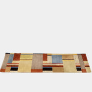 The DWR Christopher Farr Stolz 120 Multi Runner—a rectangular rug featuring a geometric pattern in beige, blue, orange, red, and brown—rests on a concrete floor against a plain white wall, evoking Gunta Stölzl’s distinctive Bauhaus-inspired aesthetic.