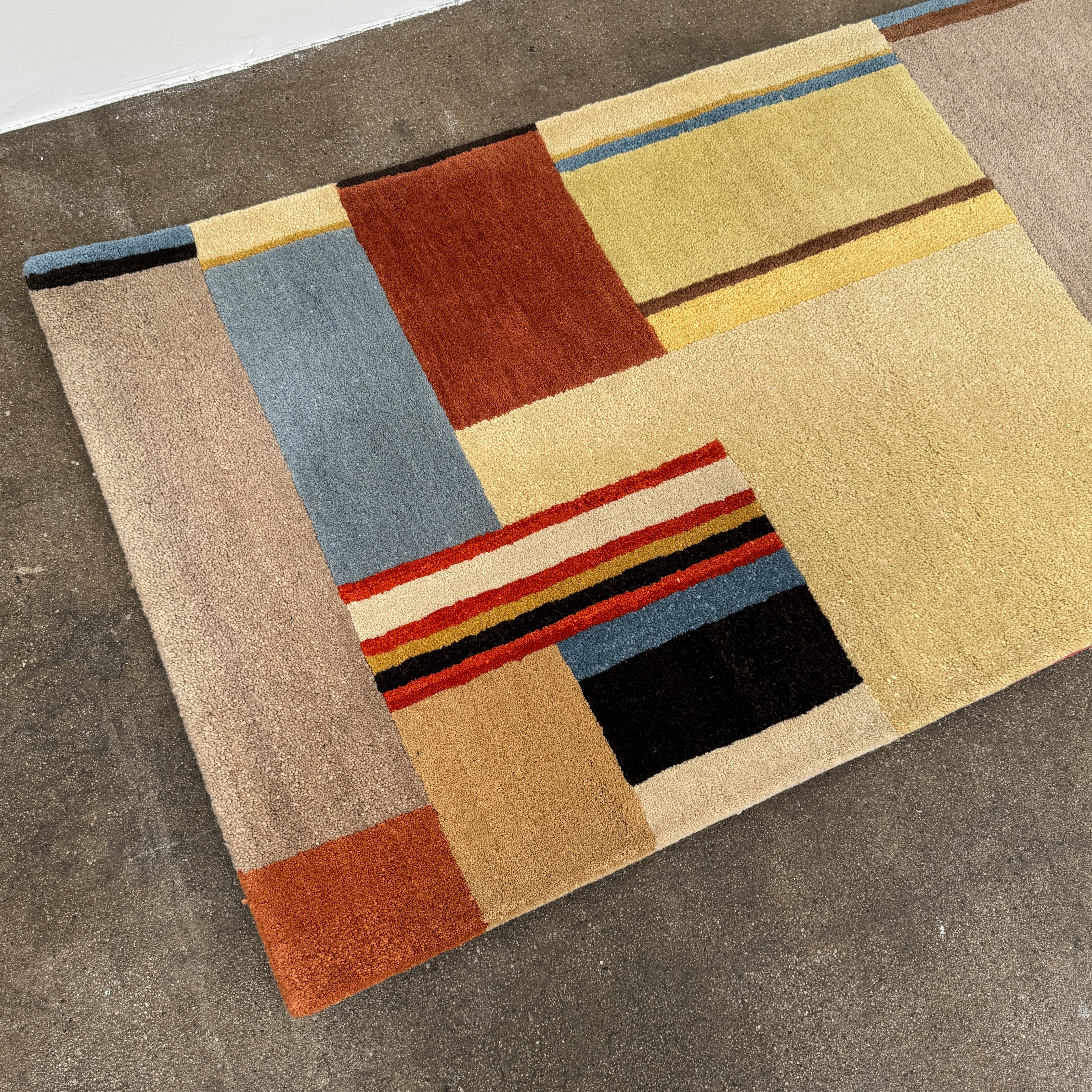 The DWR Christopher Farr Stolz 120 Multi Runner—a rectangular rug featuring a geometric pattern in beige, blue, orange, red, and brown—rests on a concrete floor against a plain white wall, evoking Gunta Stölzl’s distinctive Bauhaus-inspired aesthetic.