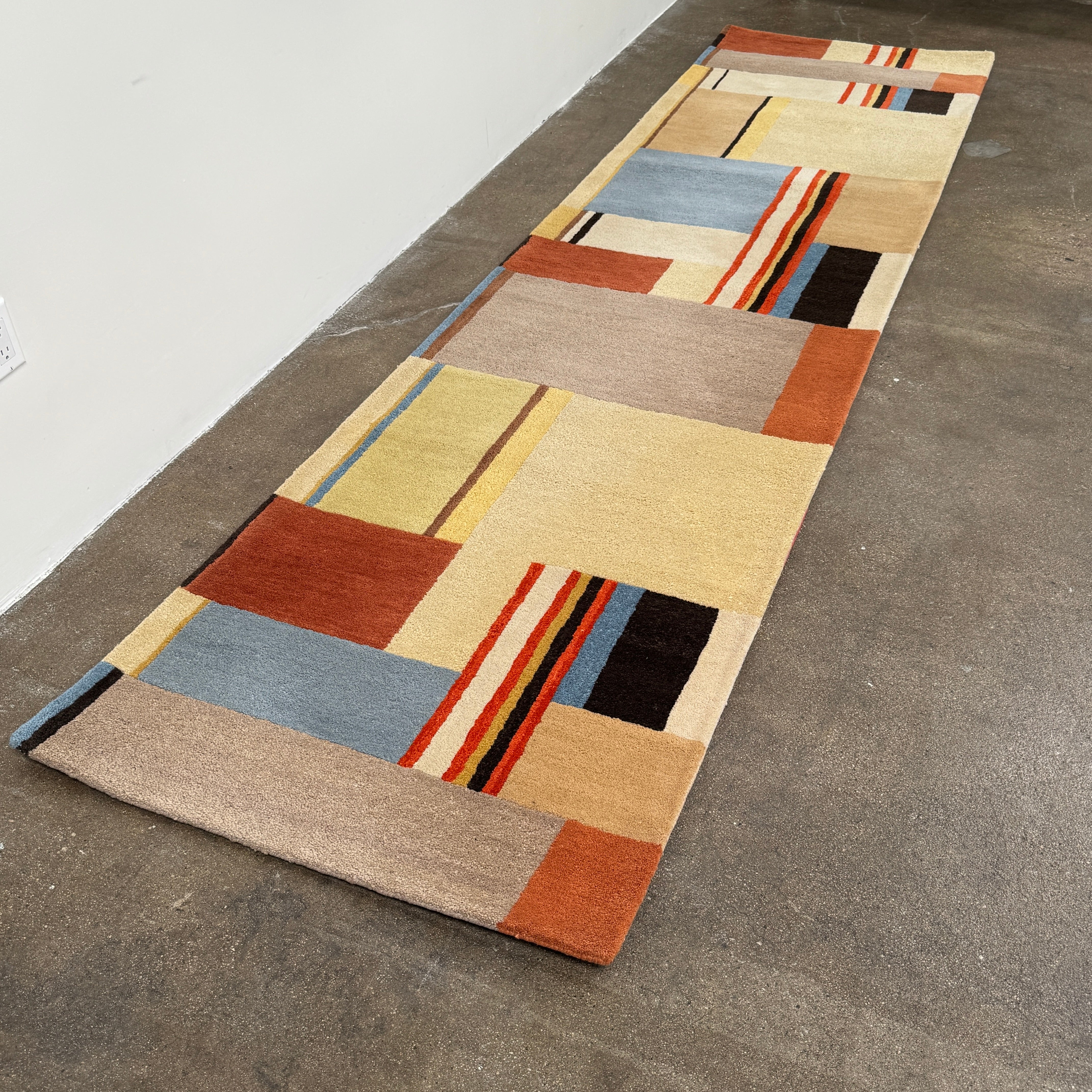 The DWR Christopher Farr Stolz 120 Multi Runner—a rectangular rug featuring a geometric pattern in beige, blue, orange, red, and brown—rests on a concrete floor against a plain white wall, evoking Gunta Stölzl’s distinctive Bauhaus-inspired aesthetic.