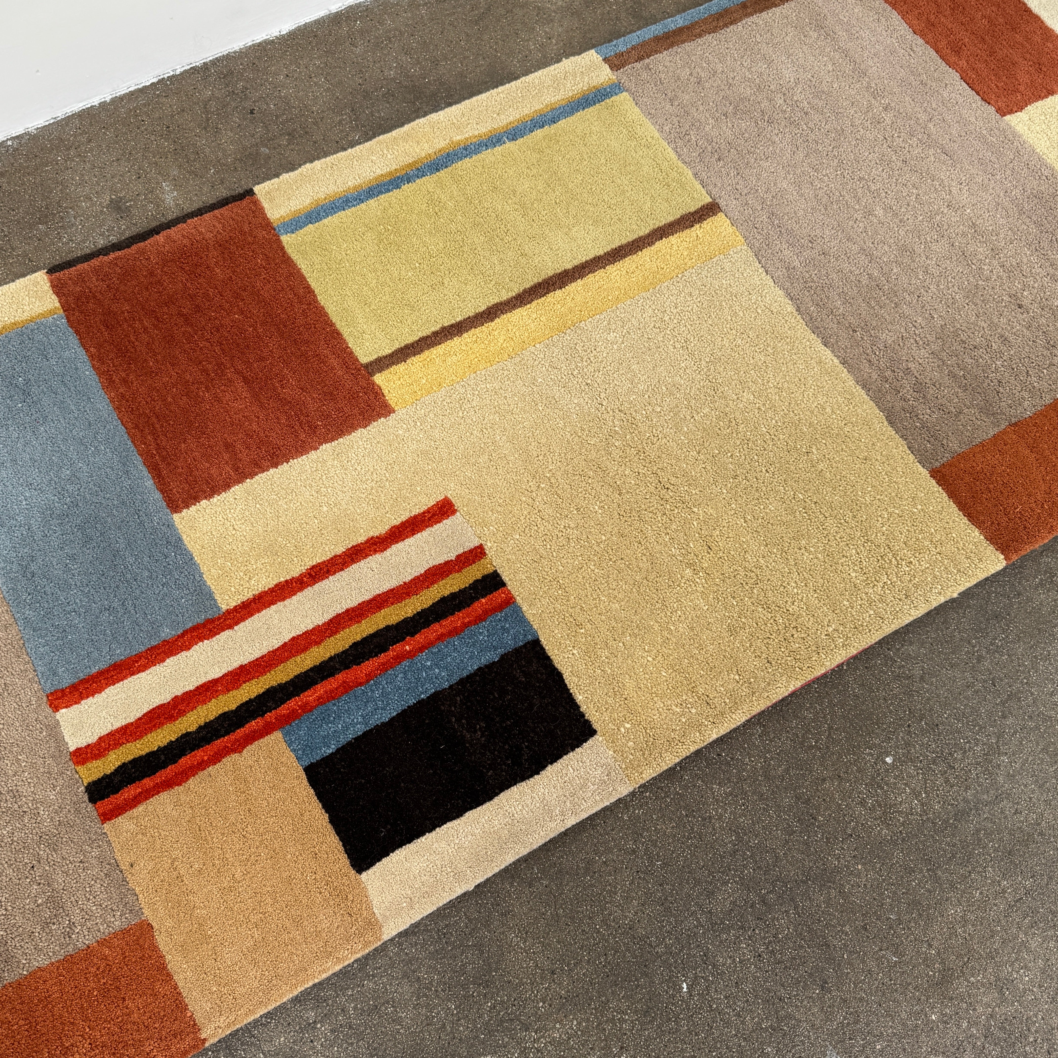 The DWR Christopher Farr Stolz 120 Multi Runner—a rectangular rug featuring a geometric pattern in beige, blue, orange, red, and brown—rests on a concrete floor against a plain white wall, evoking Gunta Stölzl’s distinctive Bauhaus-inspired aesthetic.