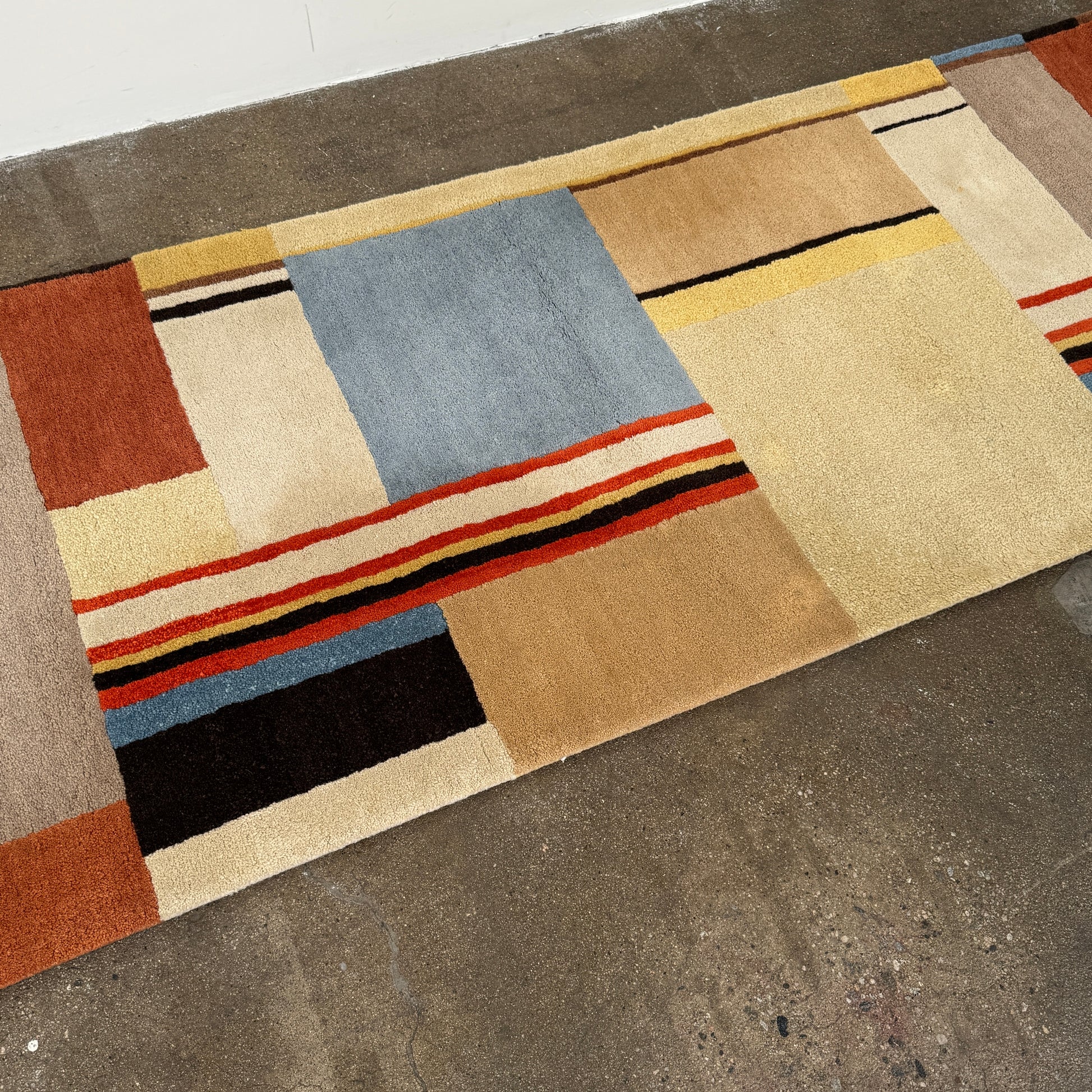 The DWR Christopher Farr Stolz 120 Multi Runner—a rectangular rug featuring a geometric pattern in beige, blue, orange, red, and brown—rests on a concrete floor against a plain white wall, evoking Gunta Stölzl’s distinctive Bauhaus-inspired aesthetic.