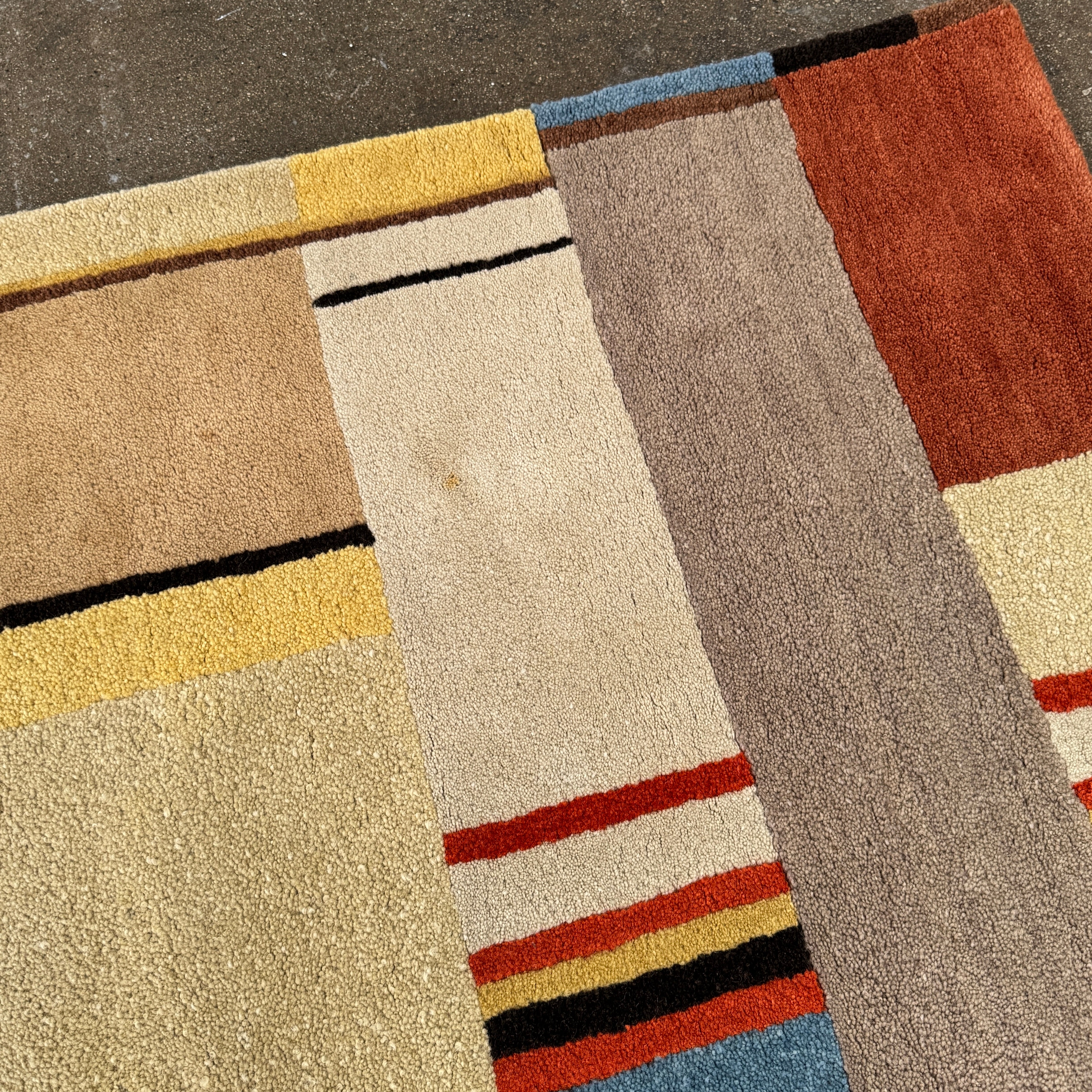 The DWR Christopher Farr Stolz 120 Multi Runner—a rectangular rug featuring a geometric pattern in beige, blue, orange, red, and brown—rests on a concrete floor against a plain white wall, evoking Gunta Stölzl’s distinctive Bauhaus-inspired aesthetic.