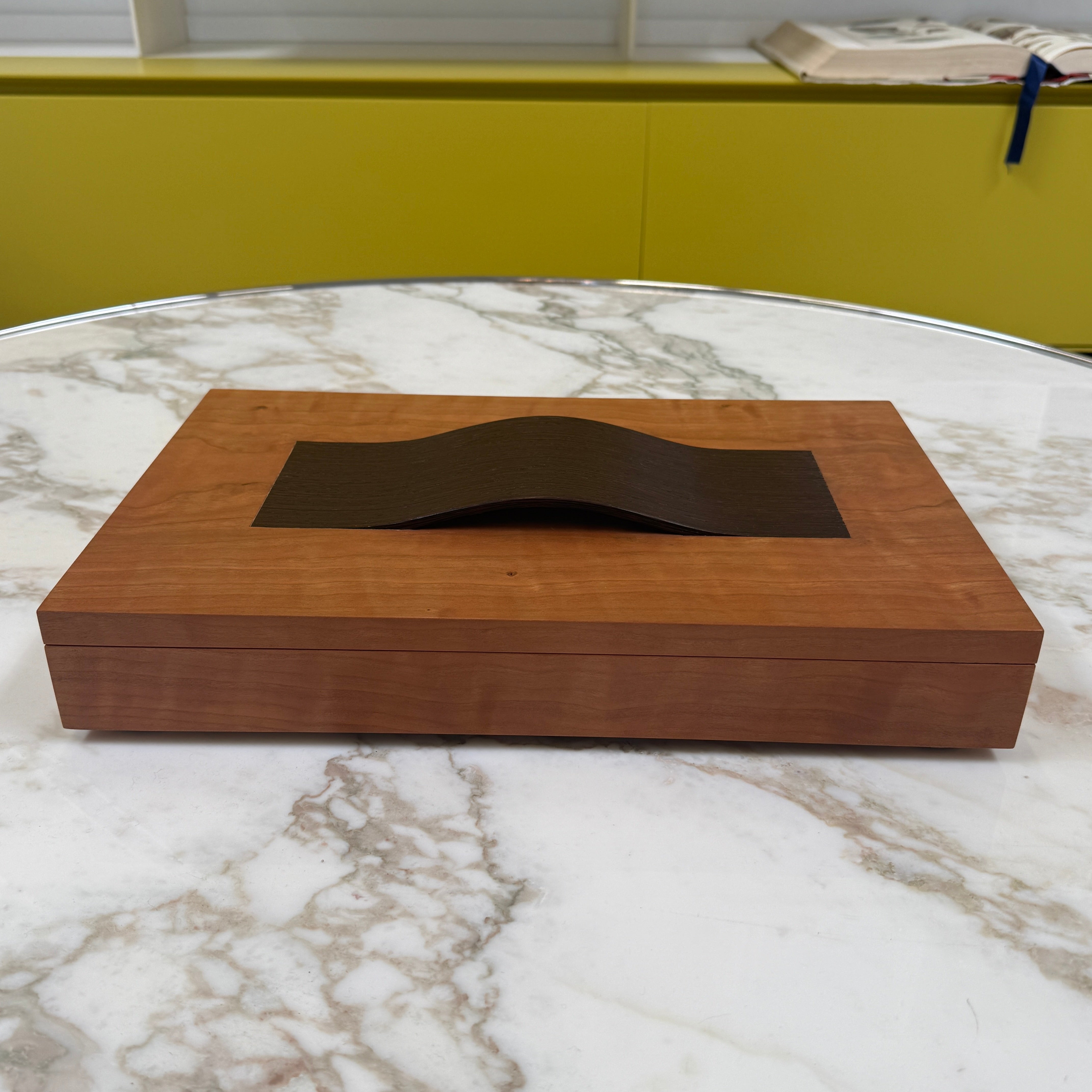 The Mike Fair Jewelry Box / Cigar Box by Mike Fair Custom Furnishings is a rectangular cherry wood box with a smooth finish and a curved, dark handle on the lid, displayed against a plain white background.