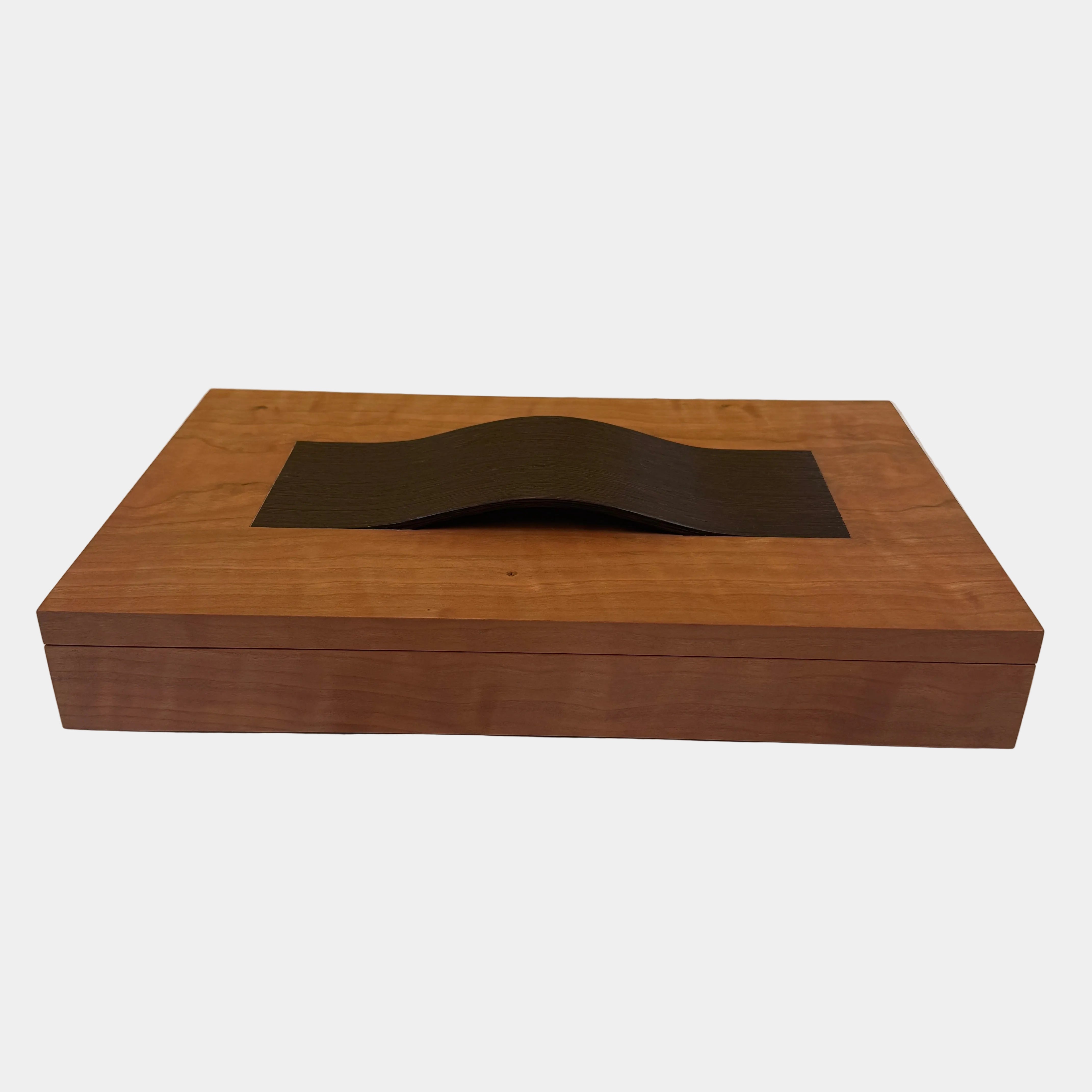 The Mike Fair Jewelry Box / Cigar Box by Mike Fair Custom Furnishings is a rectangular cherry wood box with a smooth finish and a curved, dark handle on the lid, displayed against a plain white background.