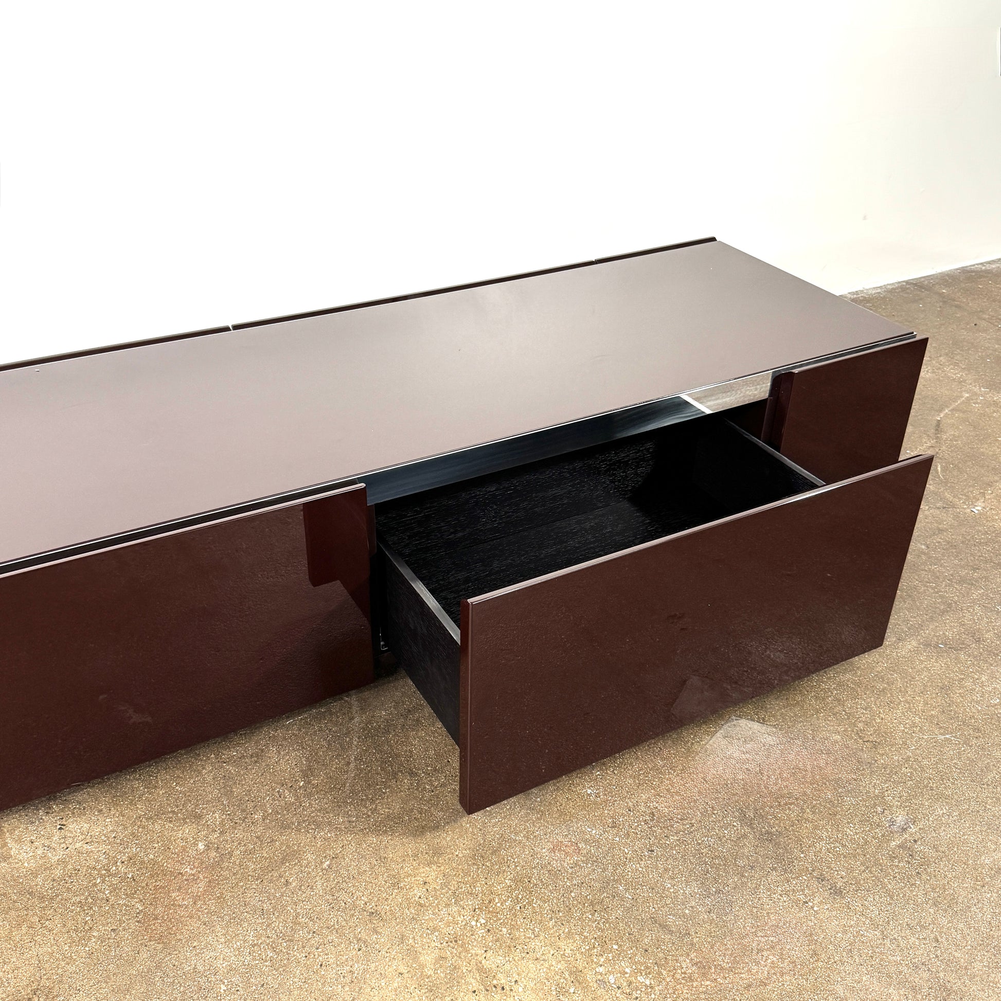 The Minotti Jason Sideboard by Minotti is a long, low-profile, dark brown modern piece with four doors, shown against a white wall on a polished concrete floor.