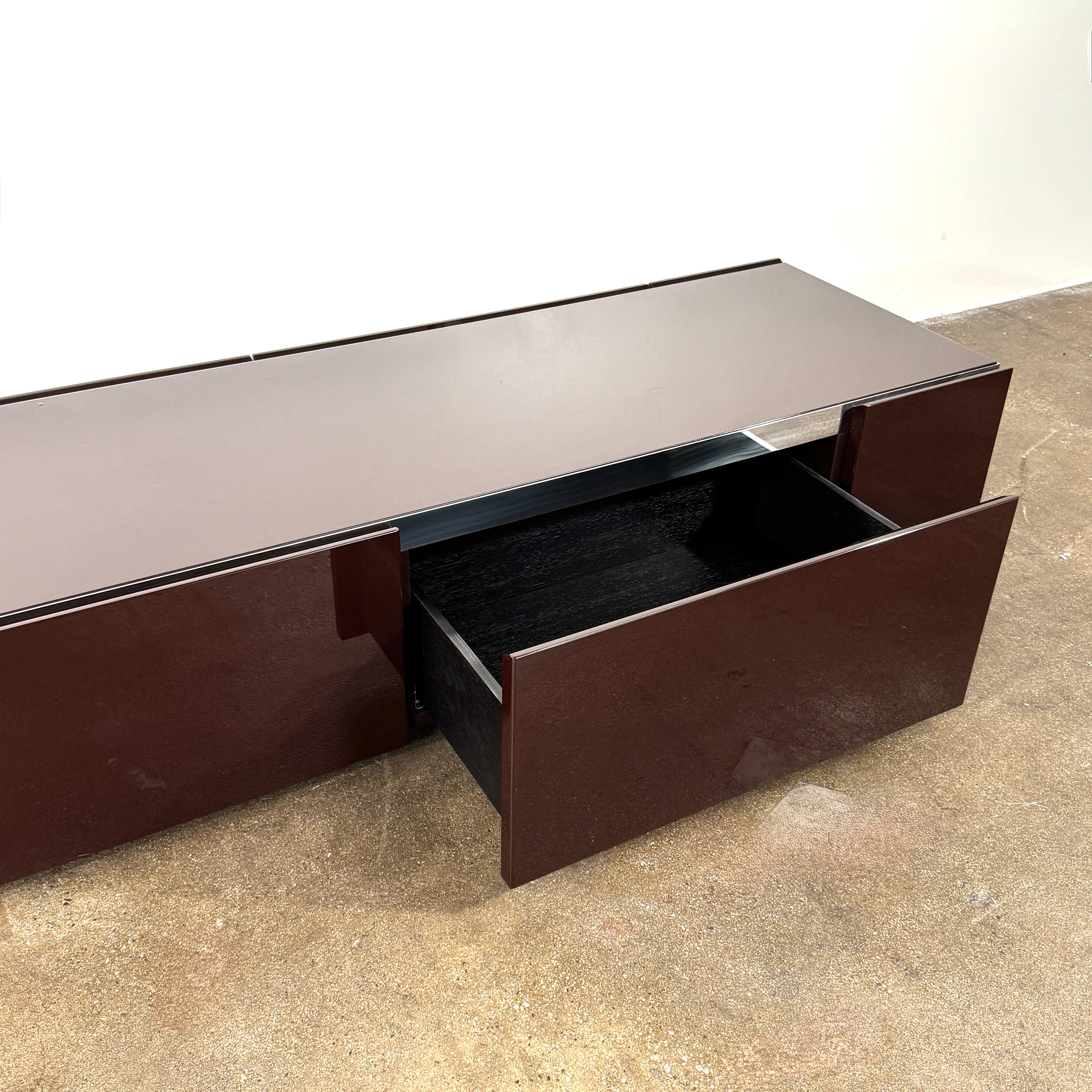 The Minotti Jason Sideboard by Minotti is a long, low-profile, dark brown modern piece with four doors, shown against a white wall on a polished concrete floor.