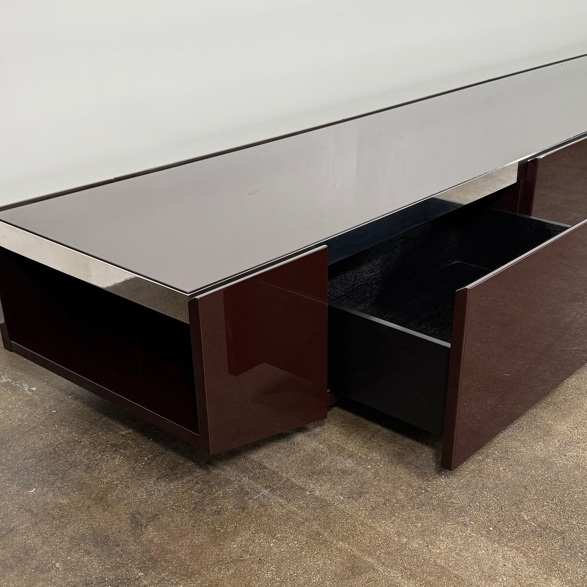 The Minotti Jason Sideboard by Minotti is a long, low-profile, dark brown modern piece with four doors, shown against a white wall on a polished concrete floor.