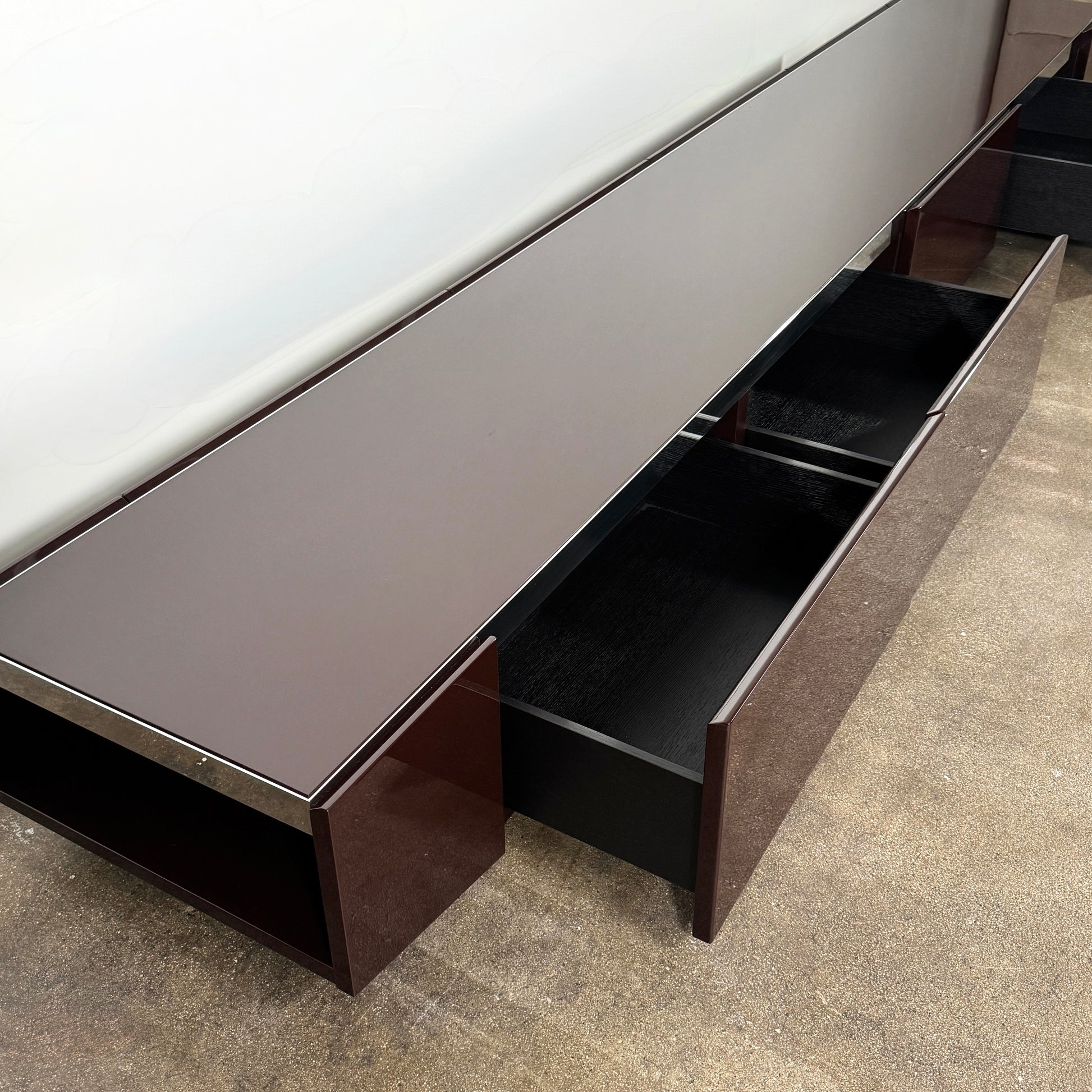 The Minotti Jason Sideboard by Minotti is a long, low-profile, dark brown modern piece with four doors, shown against a white wall on a polished concrete floor.