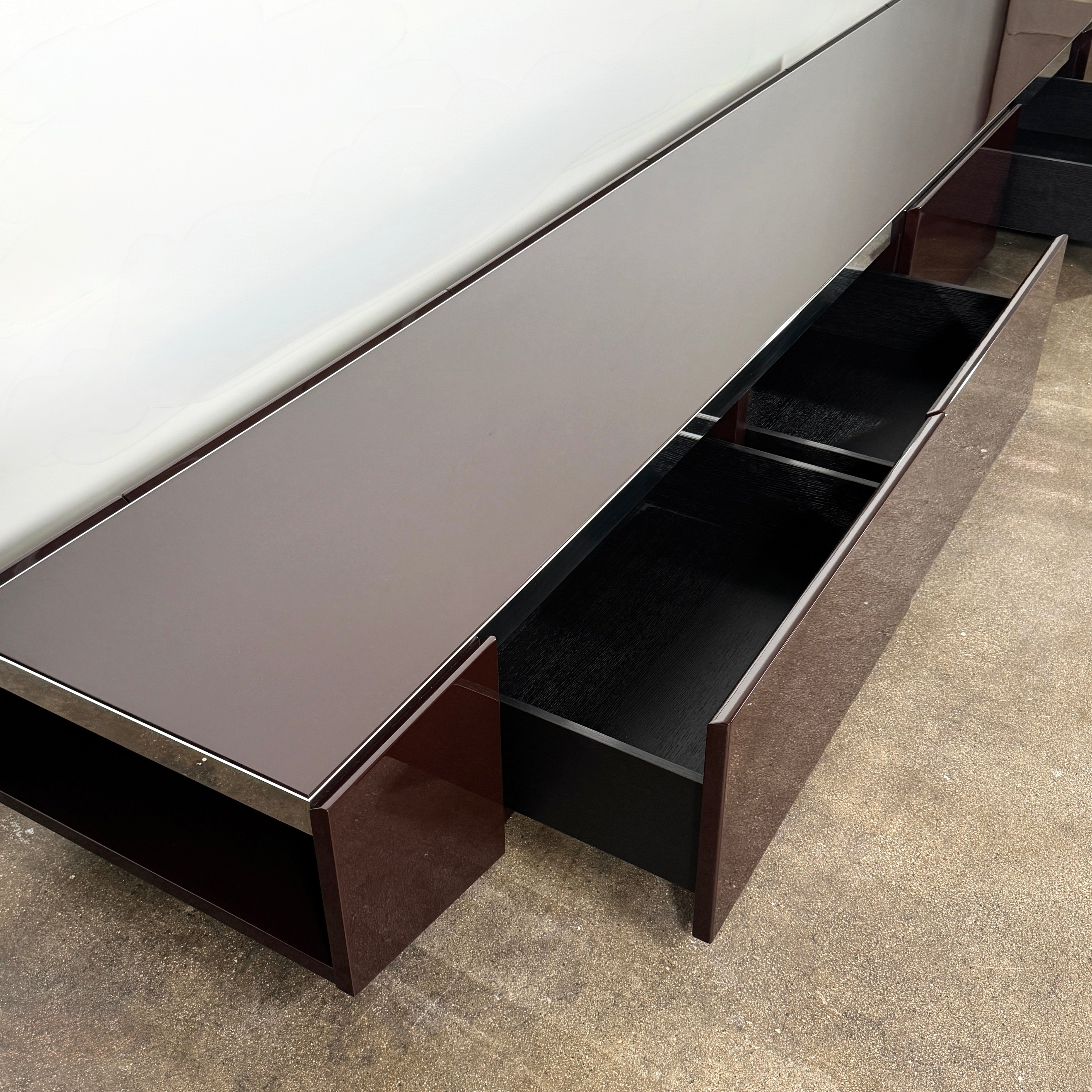 The Minotti Jason Sideboard by Minotti is a long, low-profile, dark brown modern piece with four doors, shown against a white wall on a polished concrete floor.
