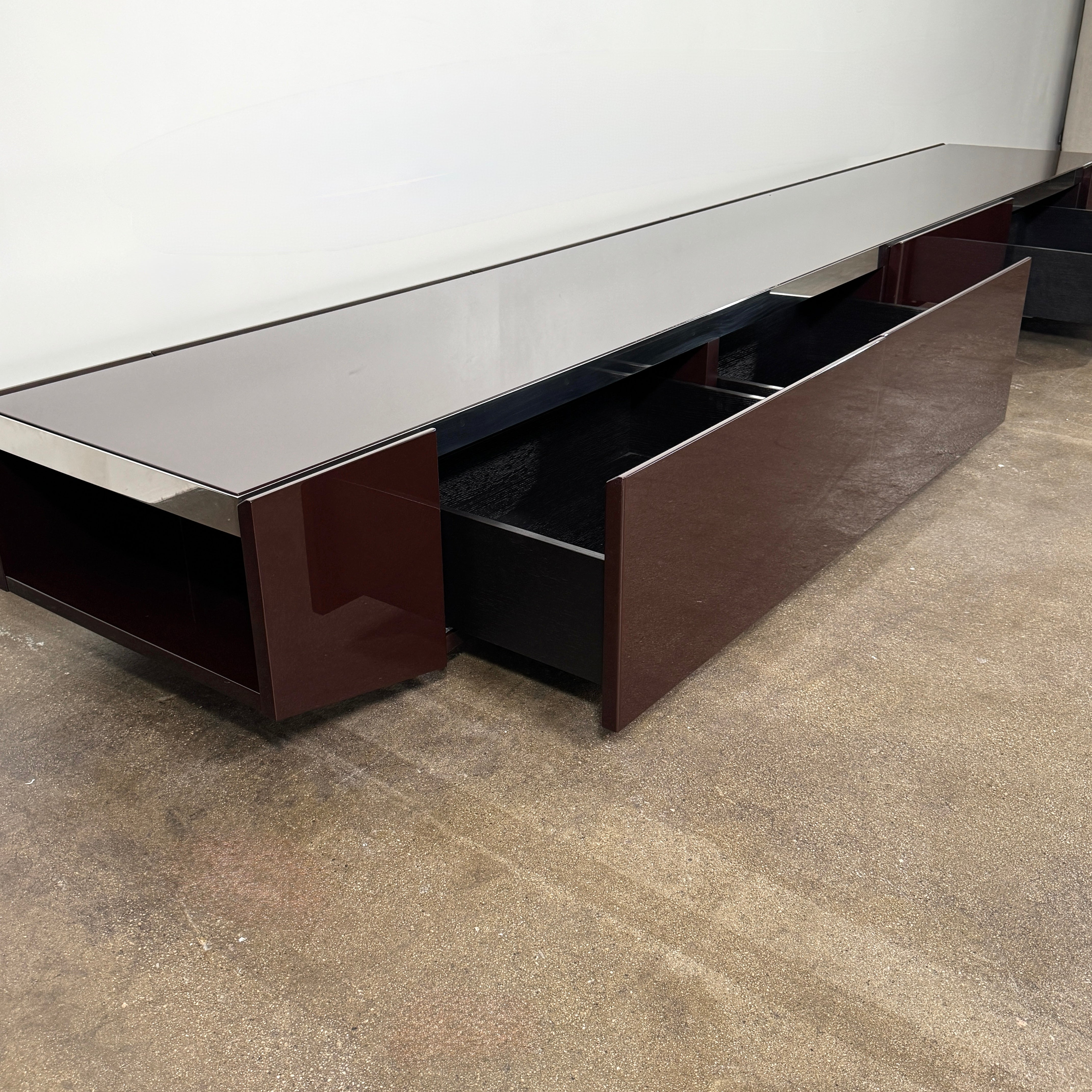 The Minotti Jason Sideboard by Minotti is a long, low-profile, dark brown modern piece with four doors, shown against a white wall on a polished concrete floor.