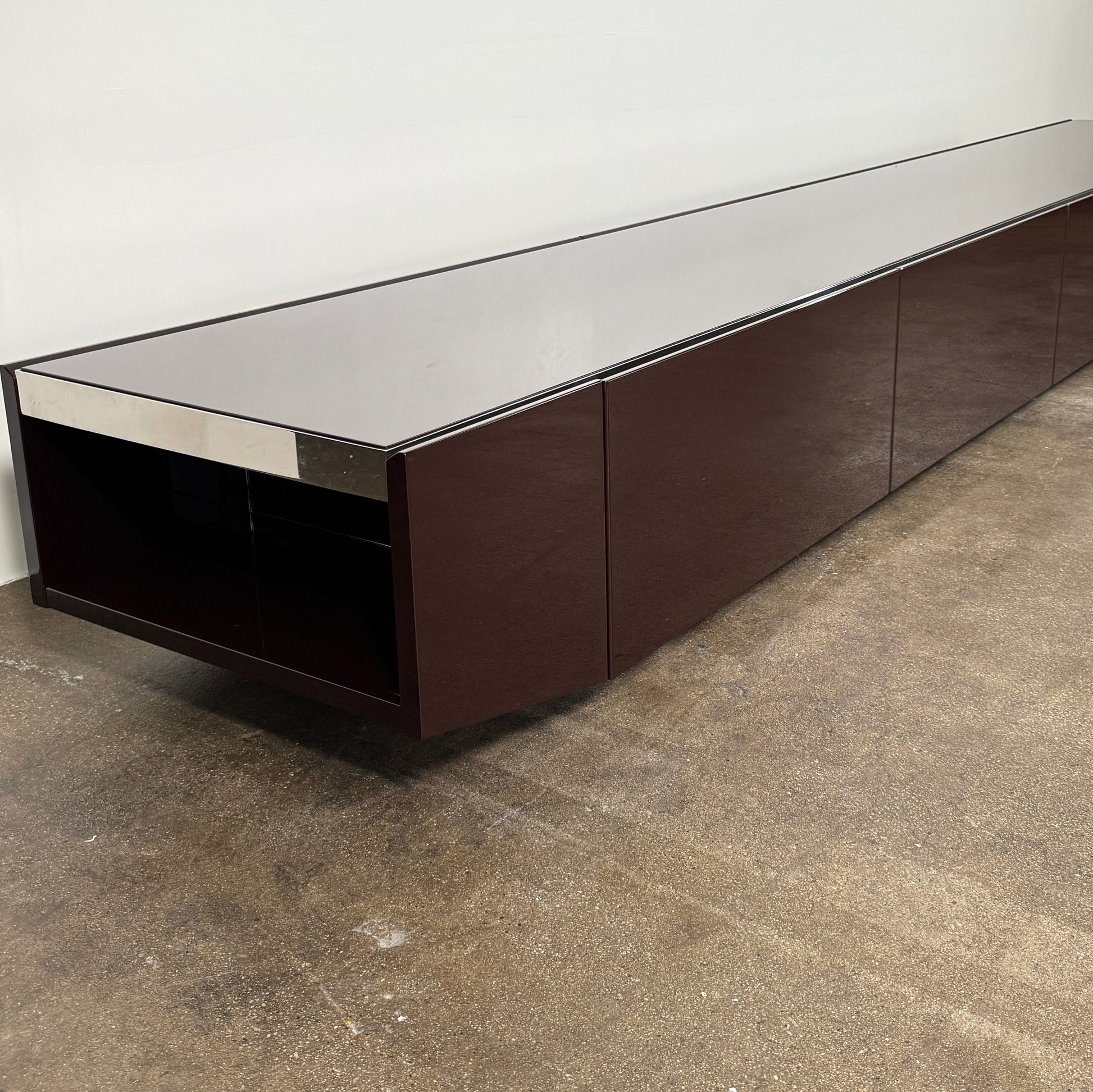 The Minotti Jason Sideboard by Minotti is a long, low-profile, dark brown modern piece with four doors, shown against a white wall on a polished concrete floor.