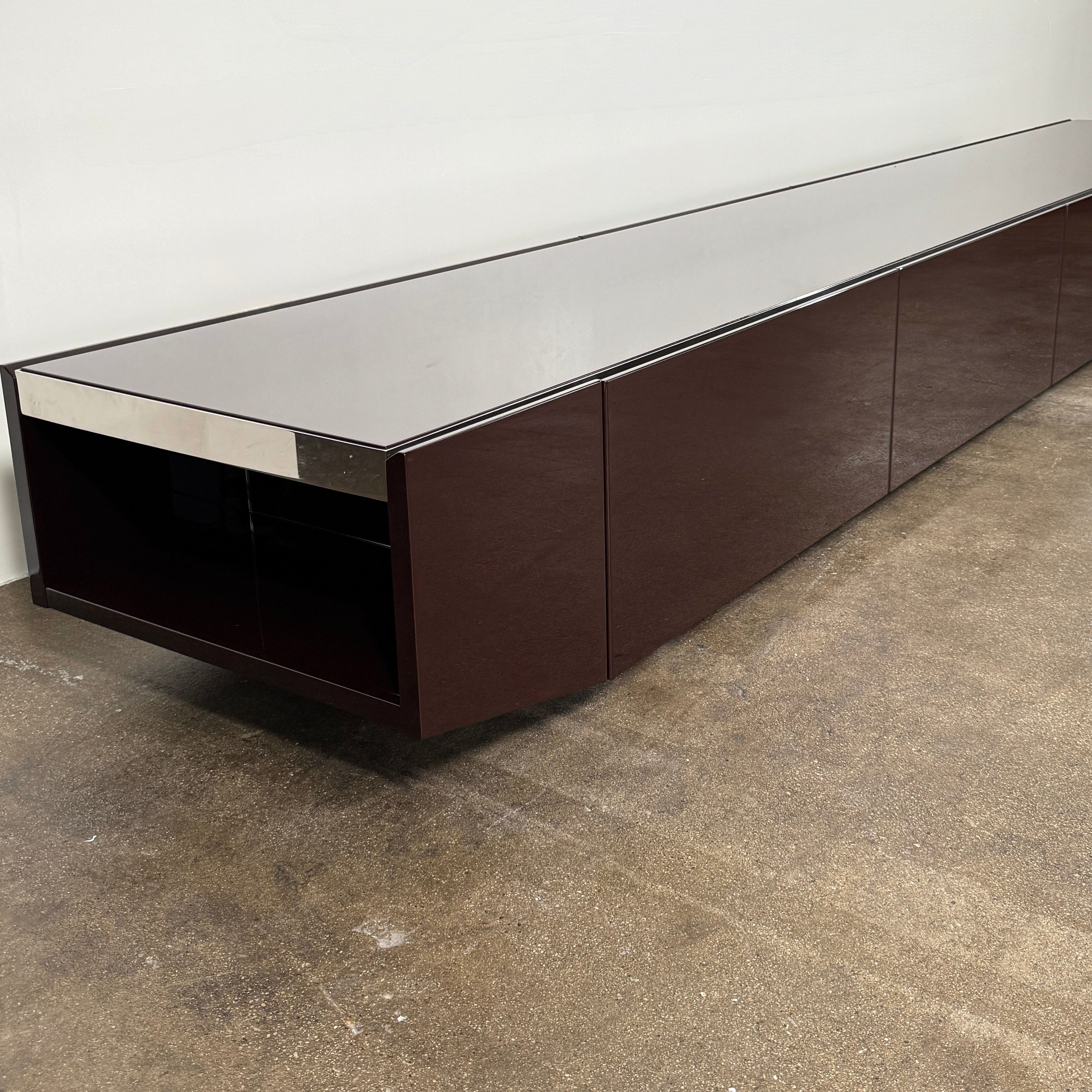 The Minotti Jason Sideboard by Minotti is a long, low-profile, dark brown modern piece with four doors, shown against a white wall on a polished concrete floor.