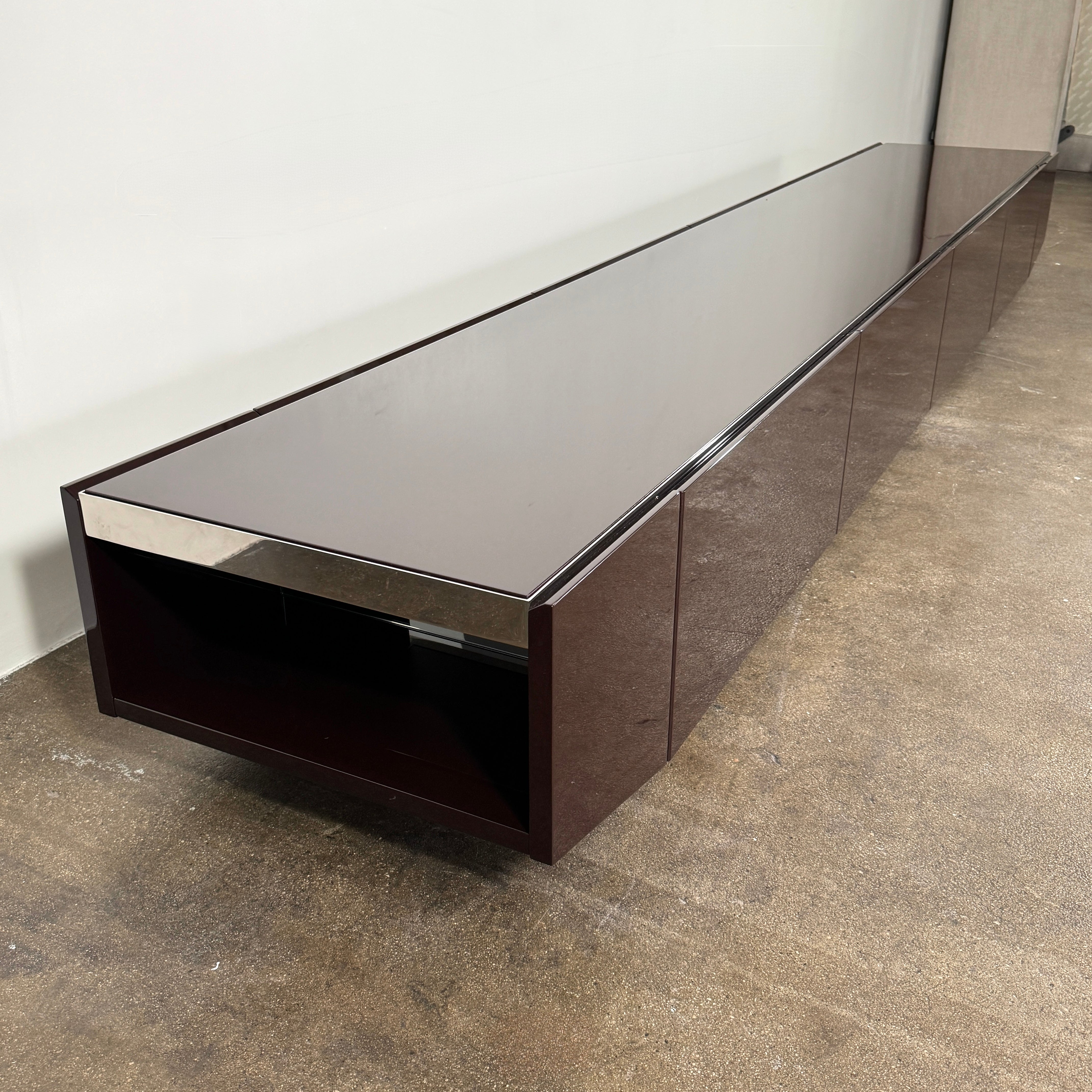 The Minotti Jason Sideboard by Minotti is a long, low-profile, dark brown modern piece with four doors, shown against a white wall on a polished concrete floor.