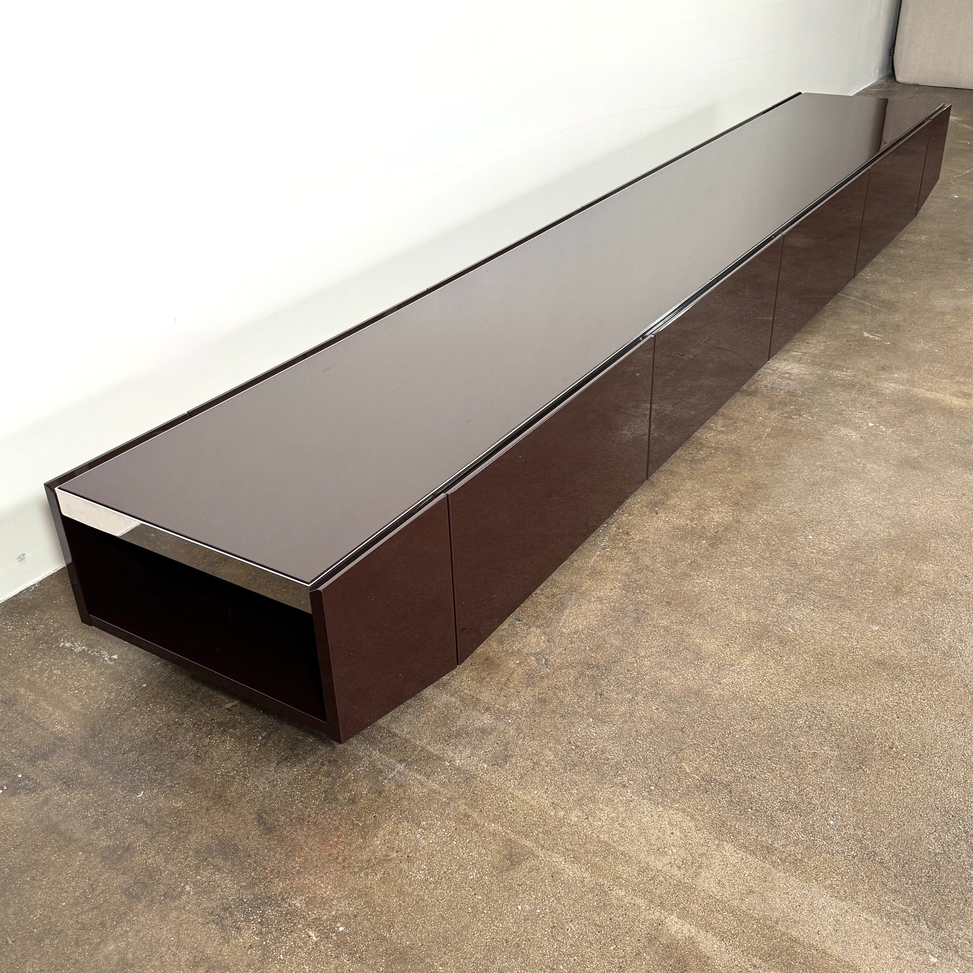 The Minotti Jason Sideboard by Minotti is a long, low-profile, dark brown modern piece with four doors, shown against a white wall on a polished concrete floor.