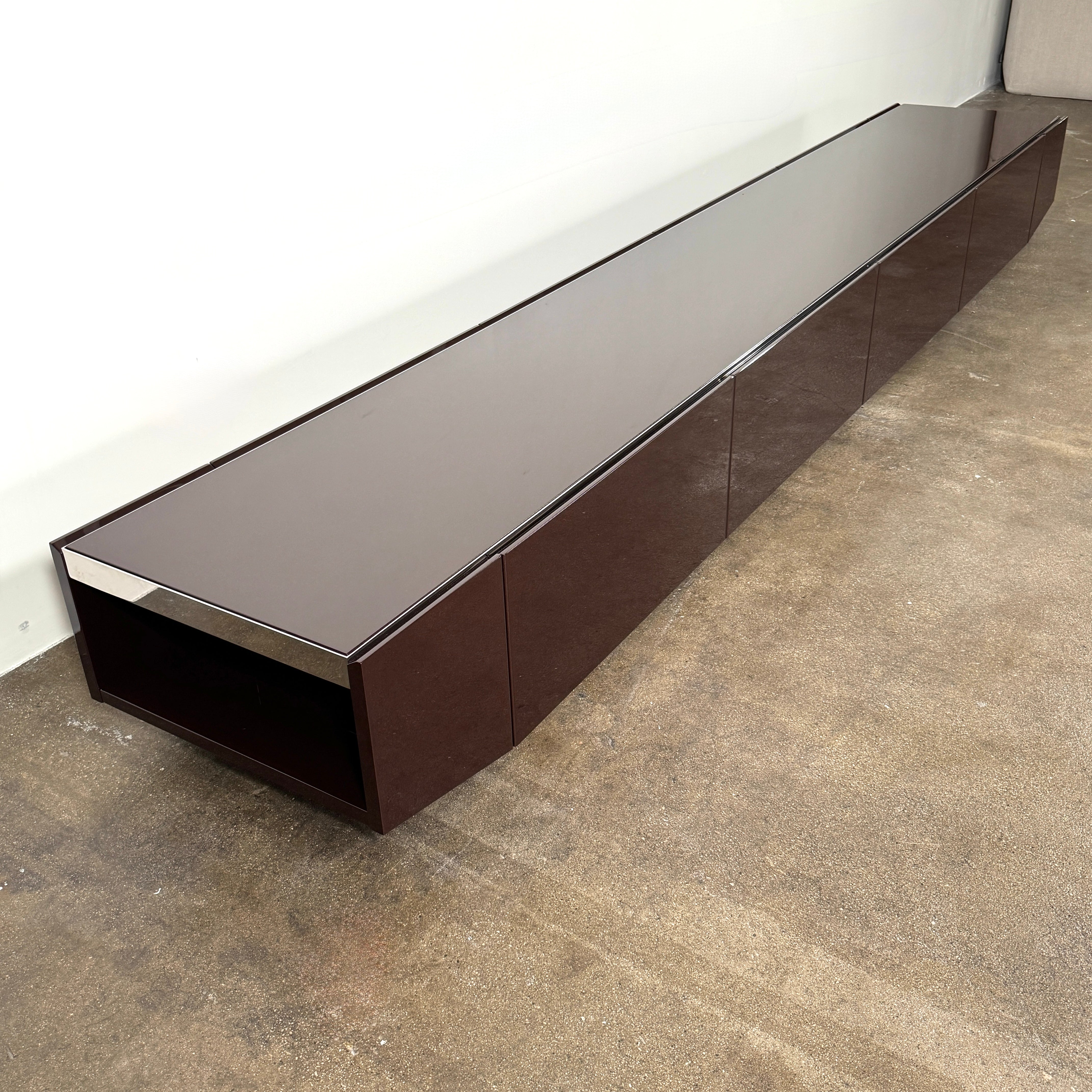 The Minotti Jason Sideboard by Minotti is a long, low-profile, dark brown modern piece with four doors, shown against a white wall on a polished concrete floor.