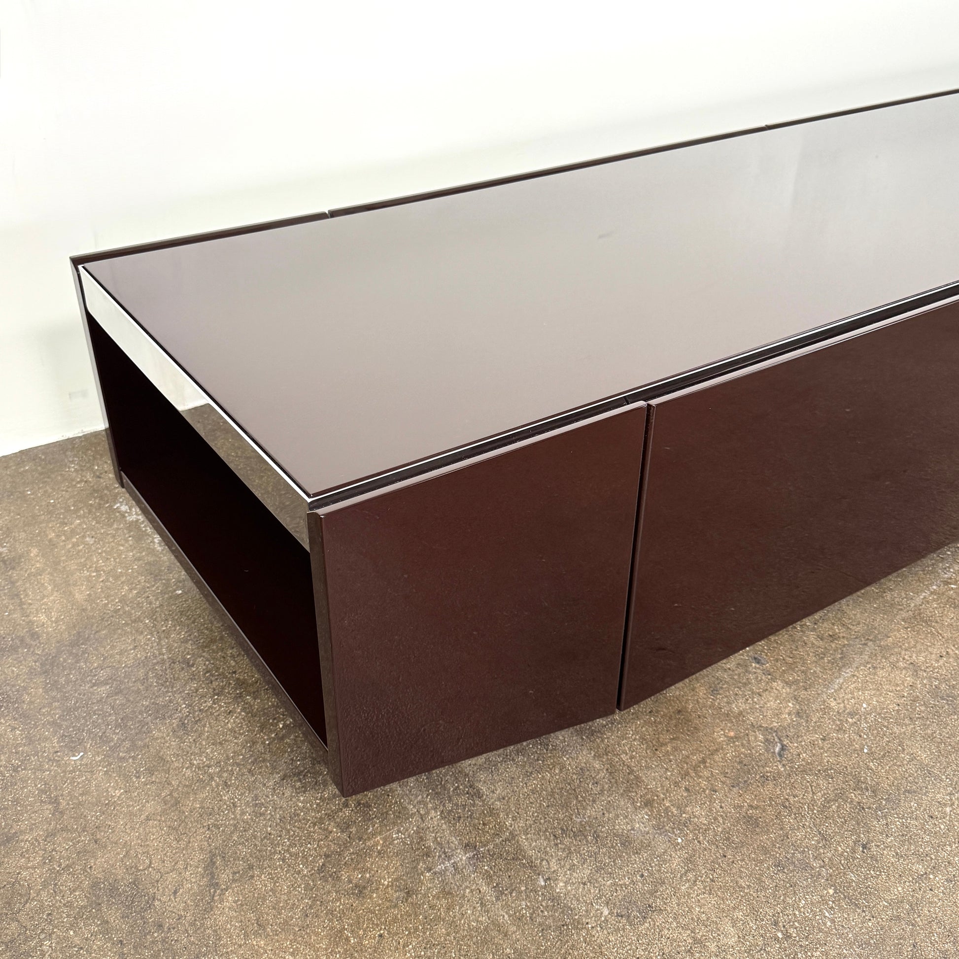 The Minotti Jason Sideboard by Minotti is a long, low-profile, dark brown modern piece with four doors, shown against a white wall on a polished concrete floor.