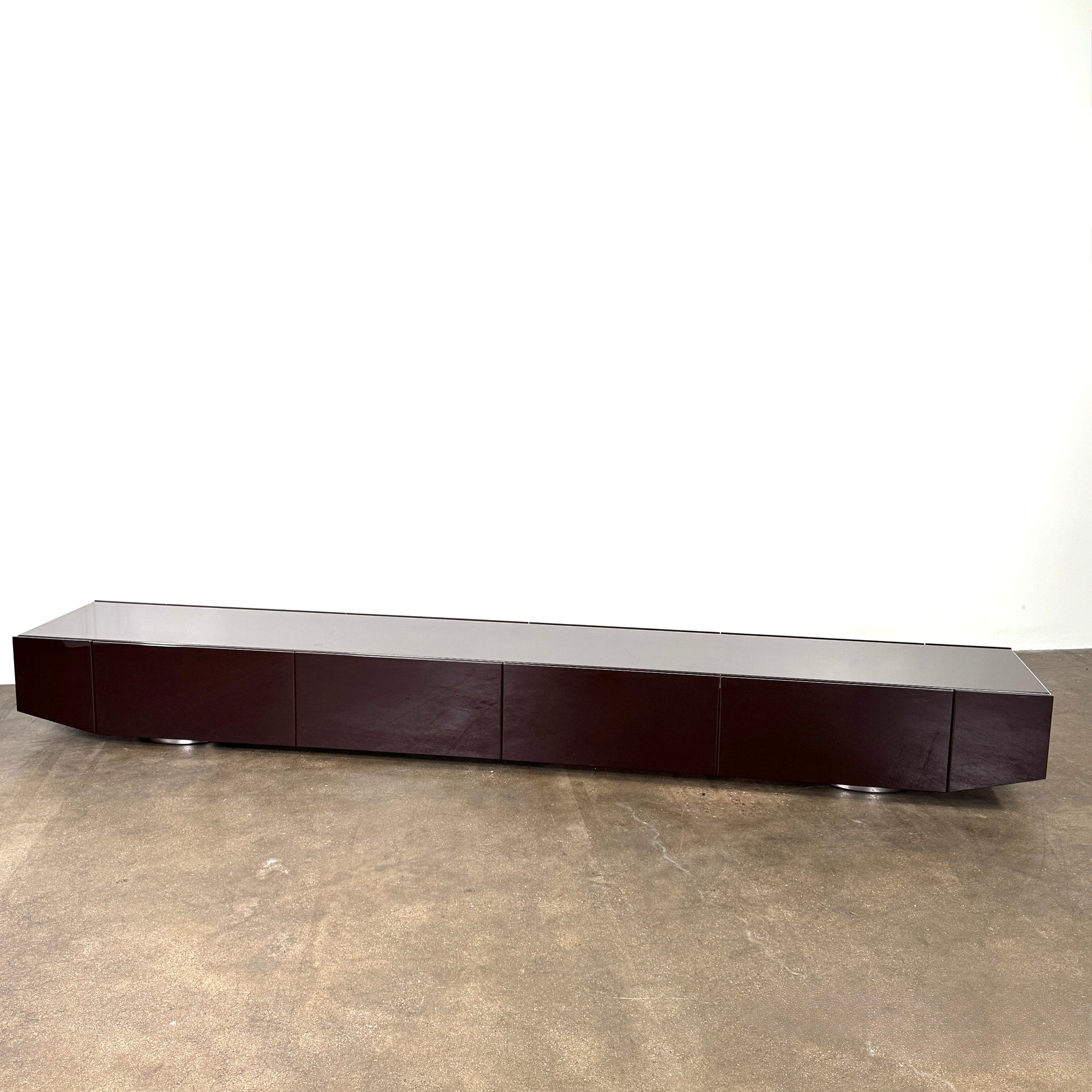 The Minotti Jason Sideboard by Minotti is a long, low-profile, dark brown modern piece with four doors, shown against a white wall on a polished concrete floor.