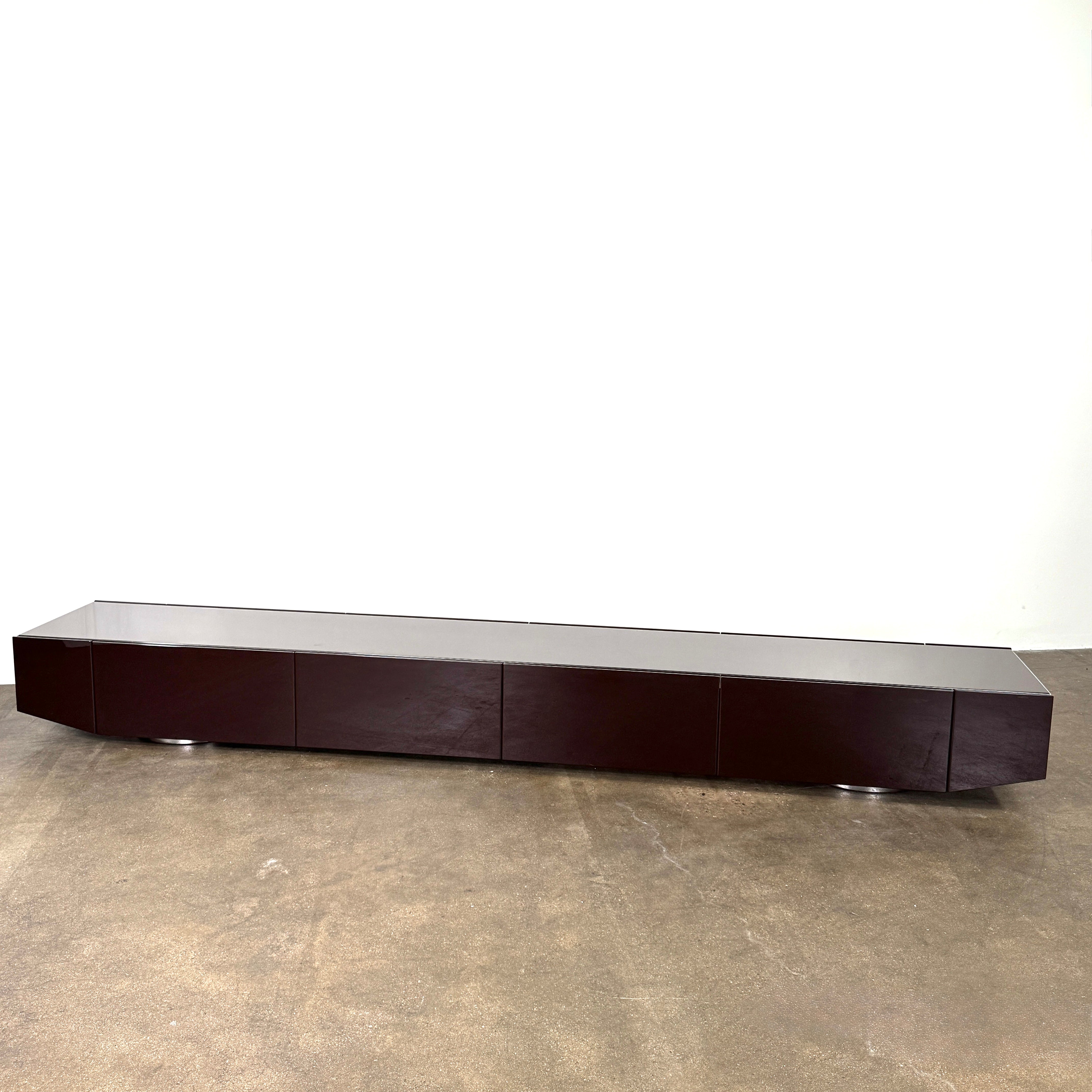 The Minotti Jason Sideboard by Minotti is a long, low-profile, dark brown modern piece with four doors, shown against a white wall on a polished concrete floor.