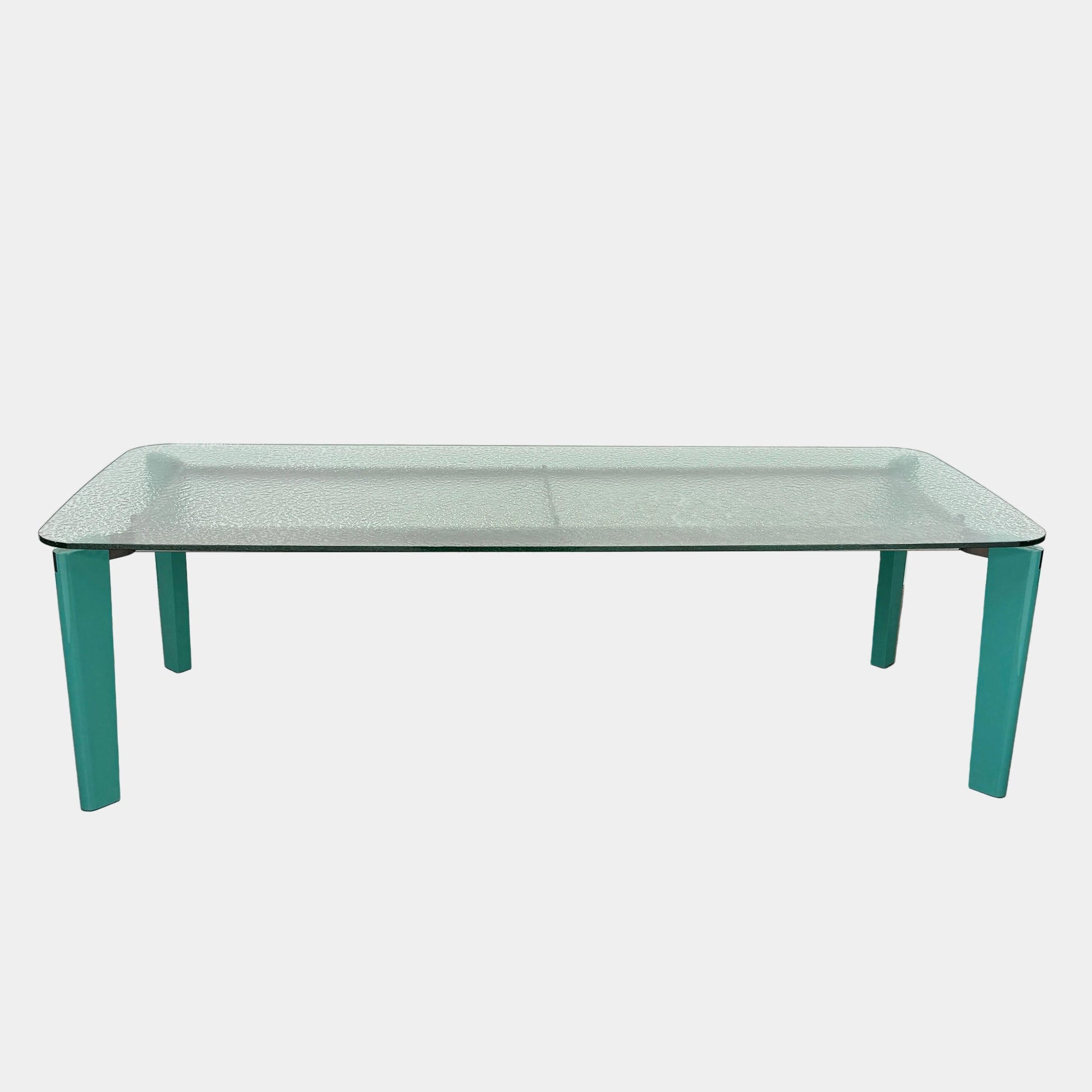 The B&B Italia Oskar Dining Table by B&B Italia features a rectangular glass top with four teal legs, displayed against a plain white background.
