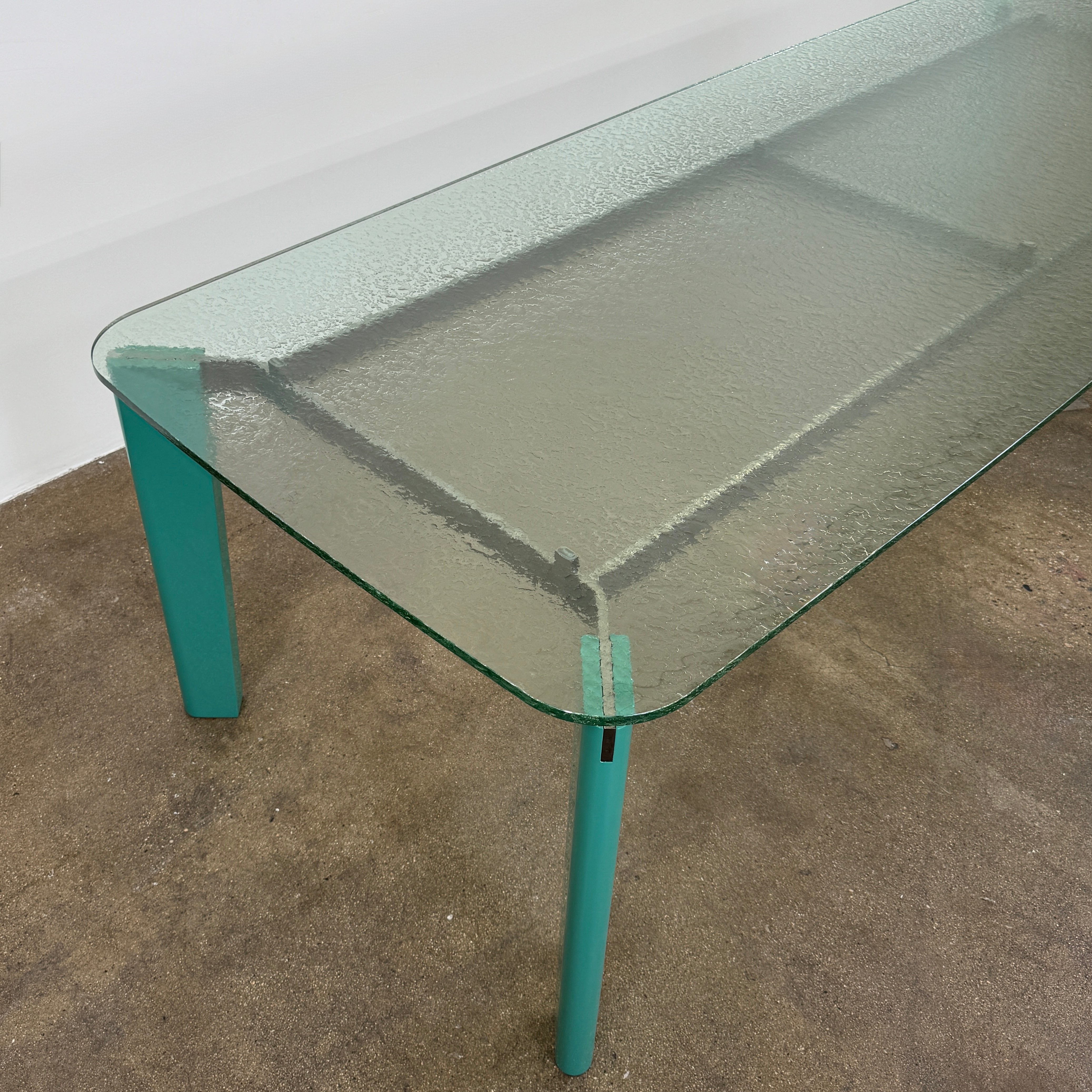 The B&B Italia Oskar Dining Table by B&B Italia features a rectangular glass top with four teal legs, displayed against a plain white background.