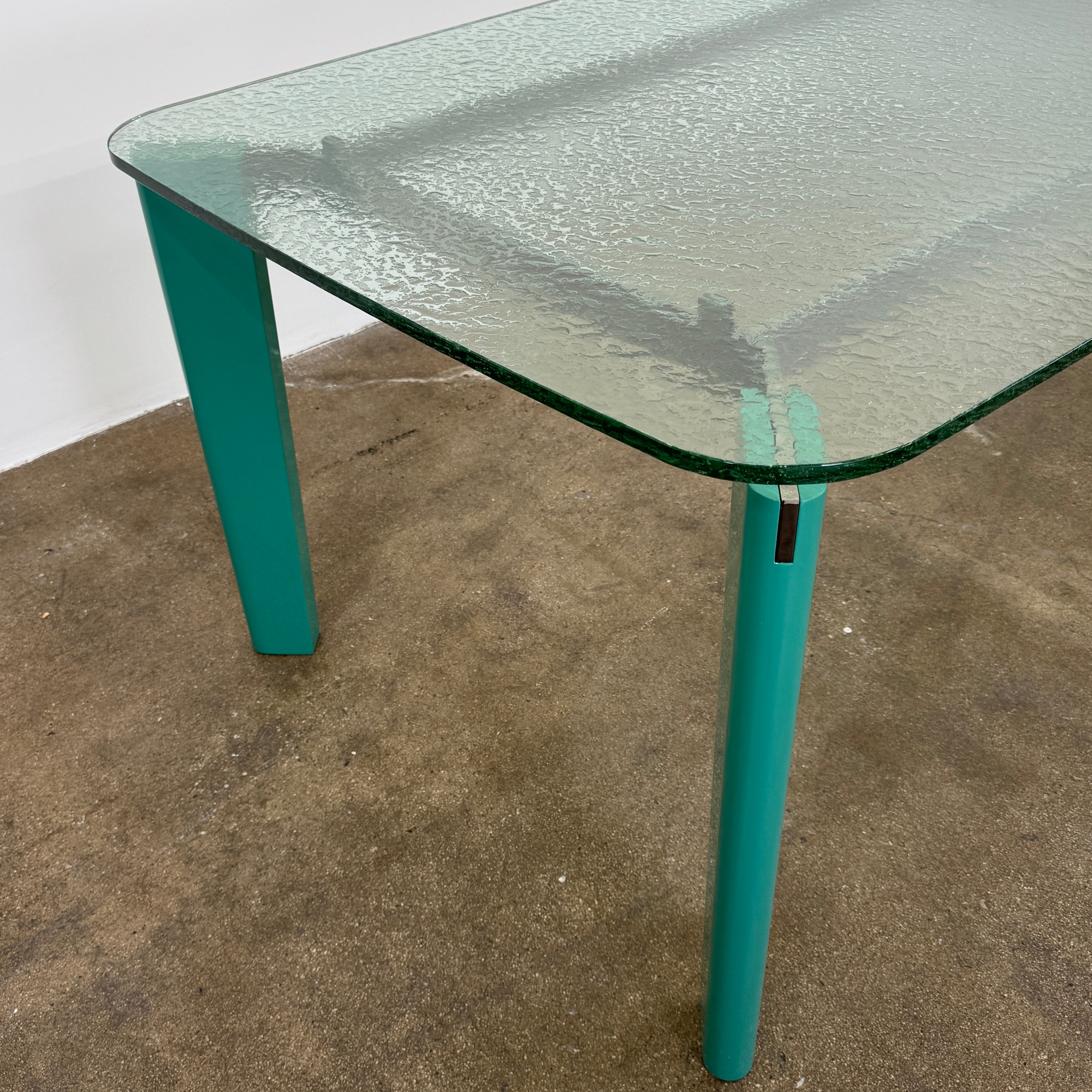 The B&B Italia Oskar Dining Table by B&B Italia features a rectangular glass top with four teal legs, displayed against a plain white background.