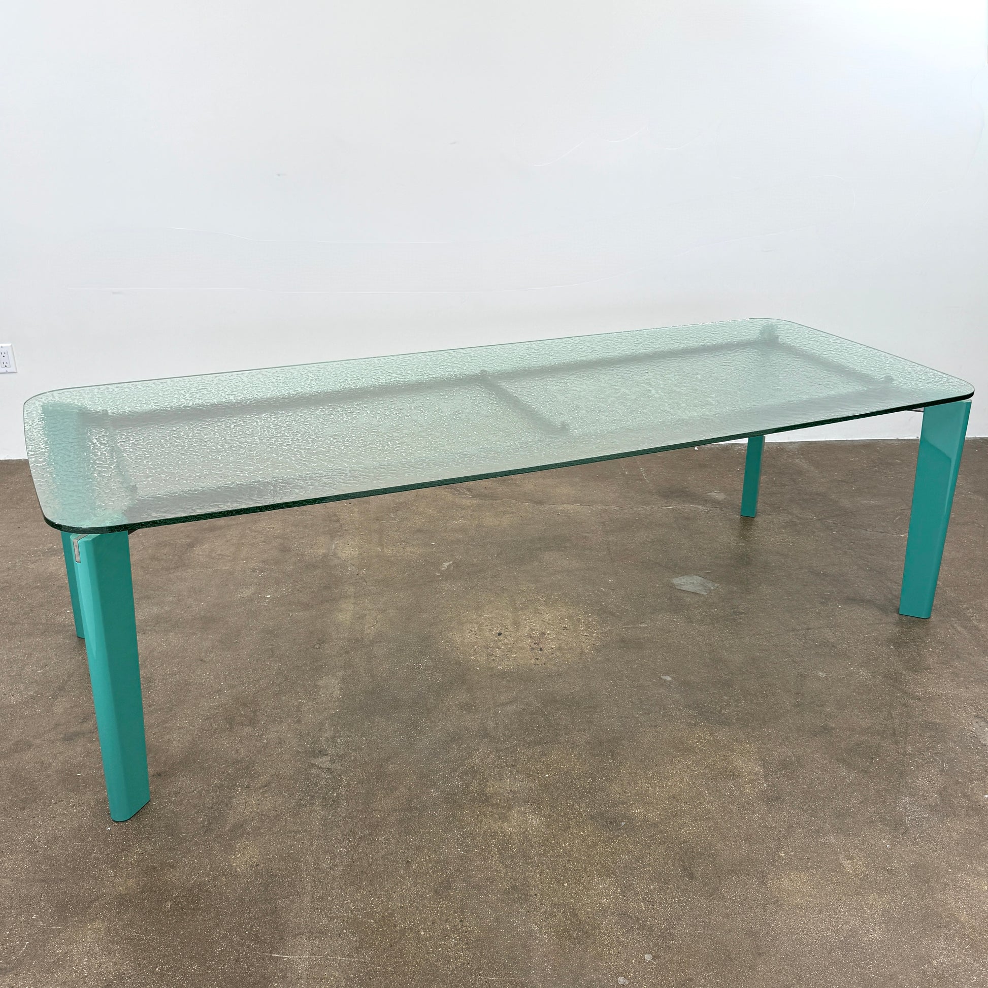 The B&B Italia Oskar Dining Table by B&B Italia features a rectangular glass top with four teal legs, displayed against a plain white background.