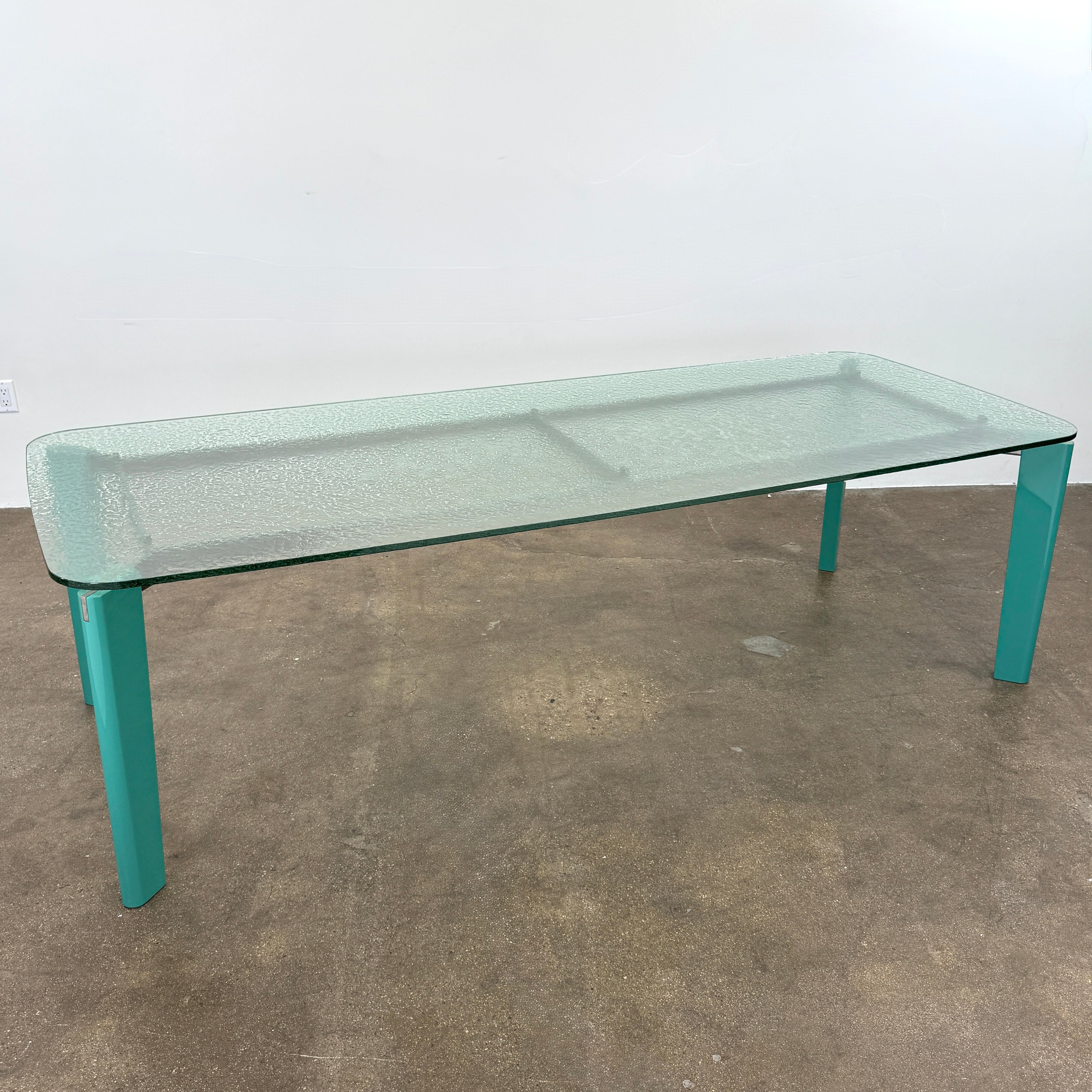 The B&B Italia Oskar Dining Table by B&B Italia features a rectangular glass top with four teal legs, displayed against a plain white background.