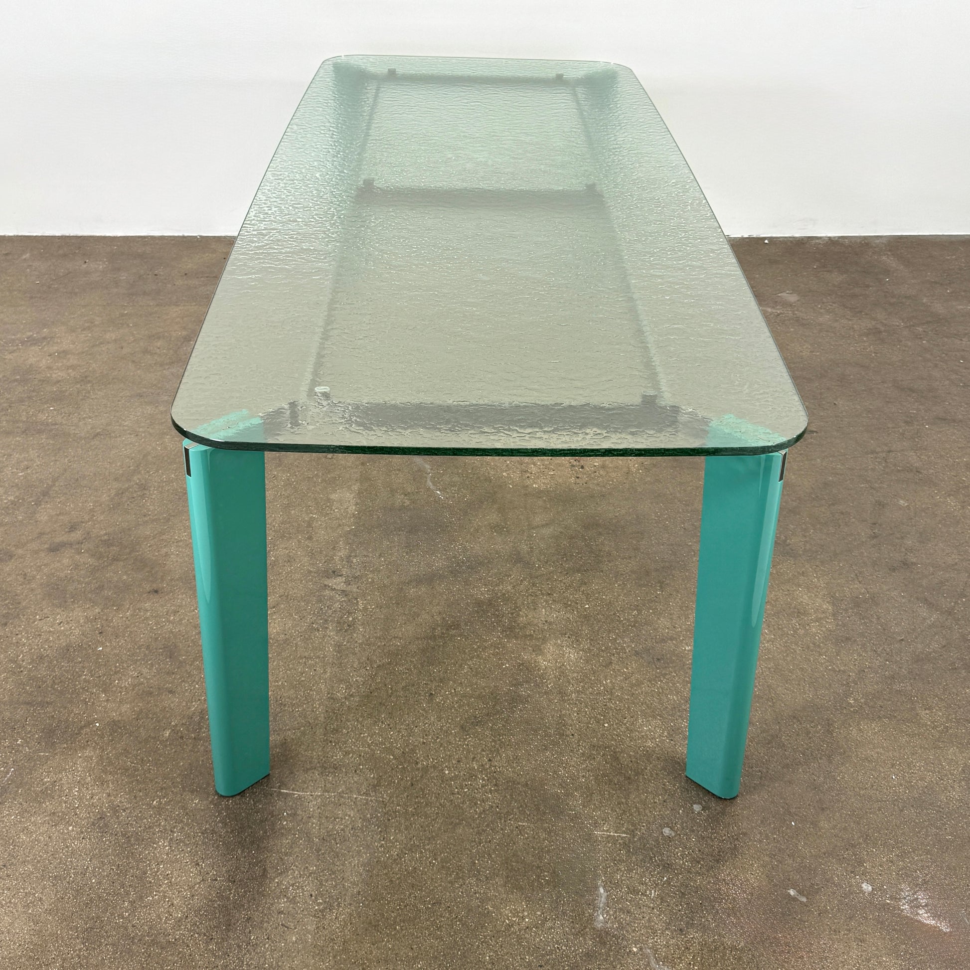 The B&B Italia Oskar Dining Table by B&B Italia features a rectangular glass top with four teal legs, displayed against a plain white background.