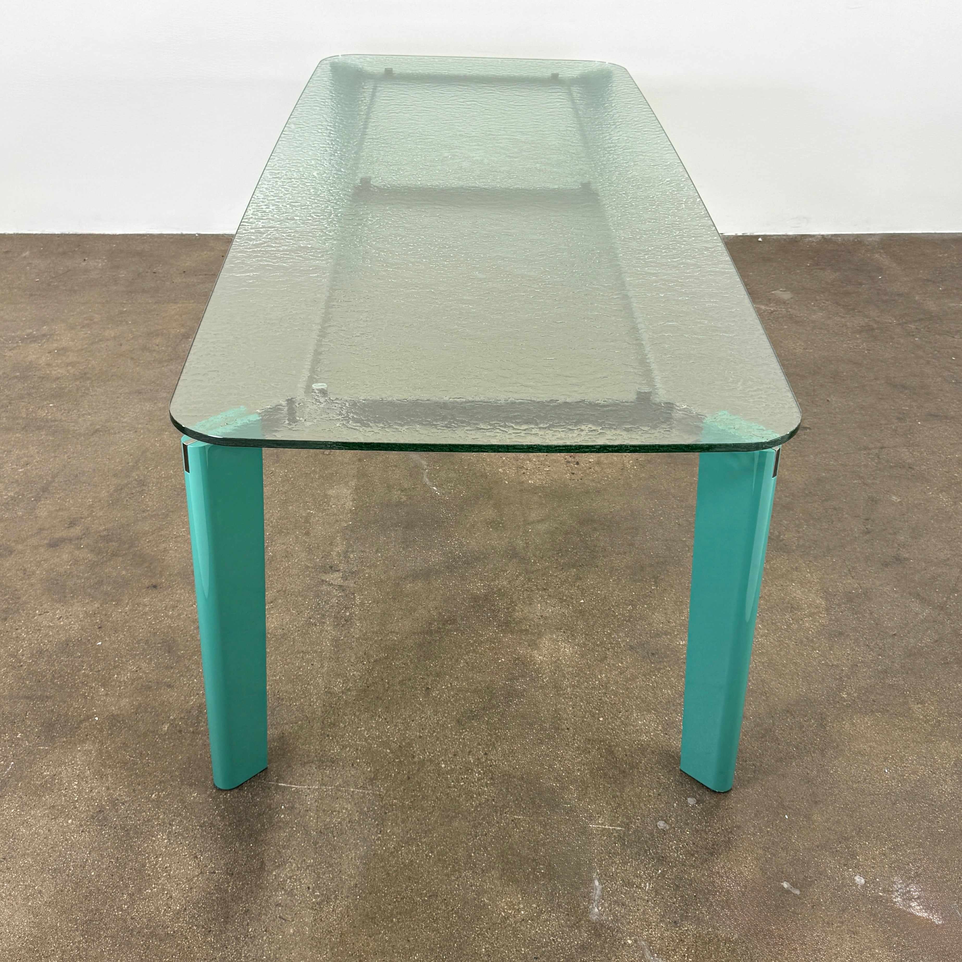 The B&B Italia Oskar Dining Table by B&B Italia features a rectangular glass top with four teal legs, displayed against a plain white background.