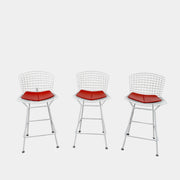 A set of three Bertoia Indoor/Outdoor Counter Stools by Vintage Modern, featuring white welded steel frames with wire backs and bold red seat cushions, lined up in a row against a simple white background—ideal for adding modern style to any indoor or outdoor space.