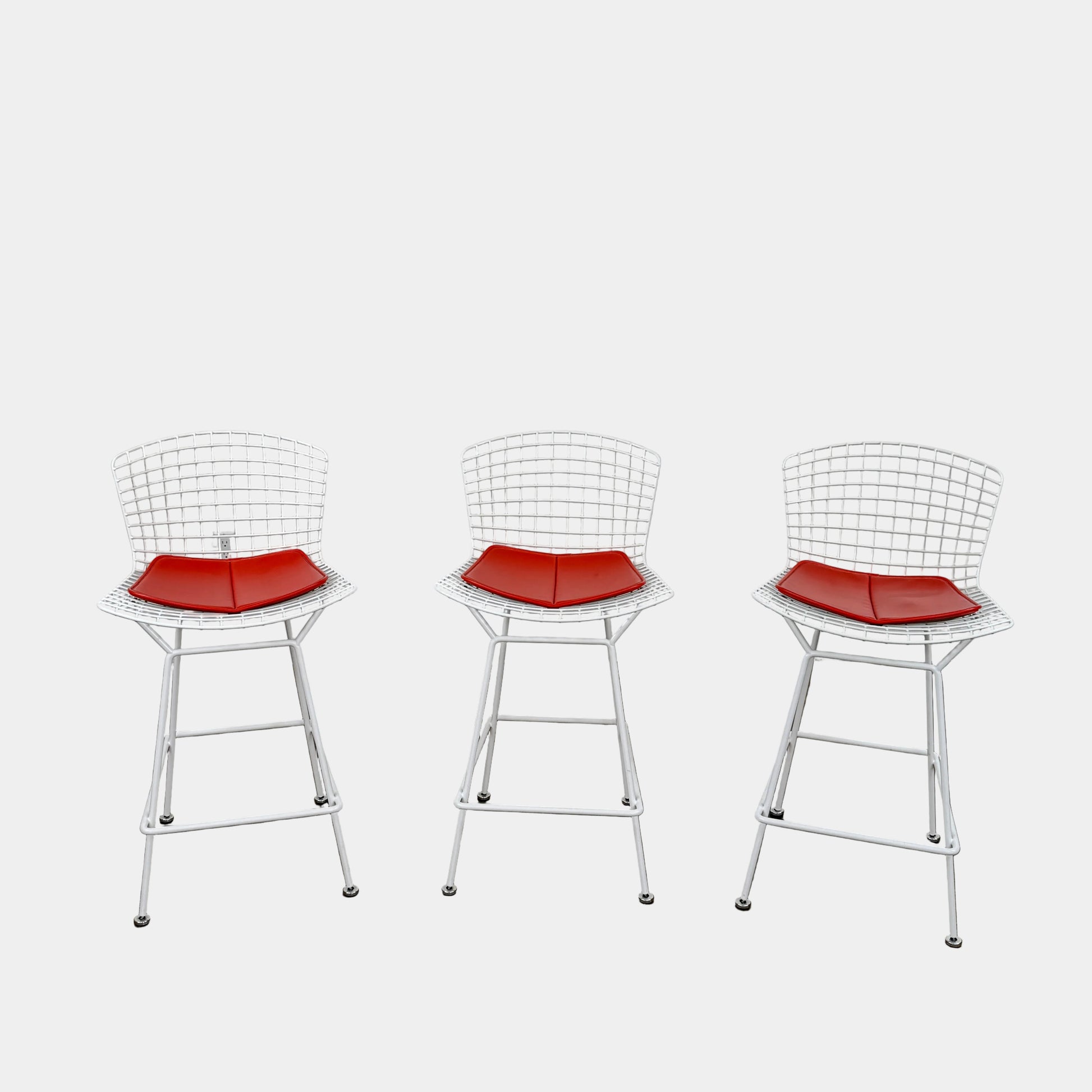 A set of three Bertoia Indoor/Outdoor Counter Stools by Vintage Modern, featuring white welded steel frames with wire backs and bold red seat cushions, lined up in a row against a simple white background—ideal for adding modern style to any indoor or outdoor space.