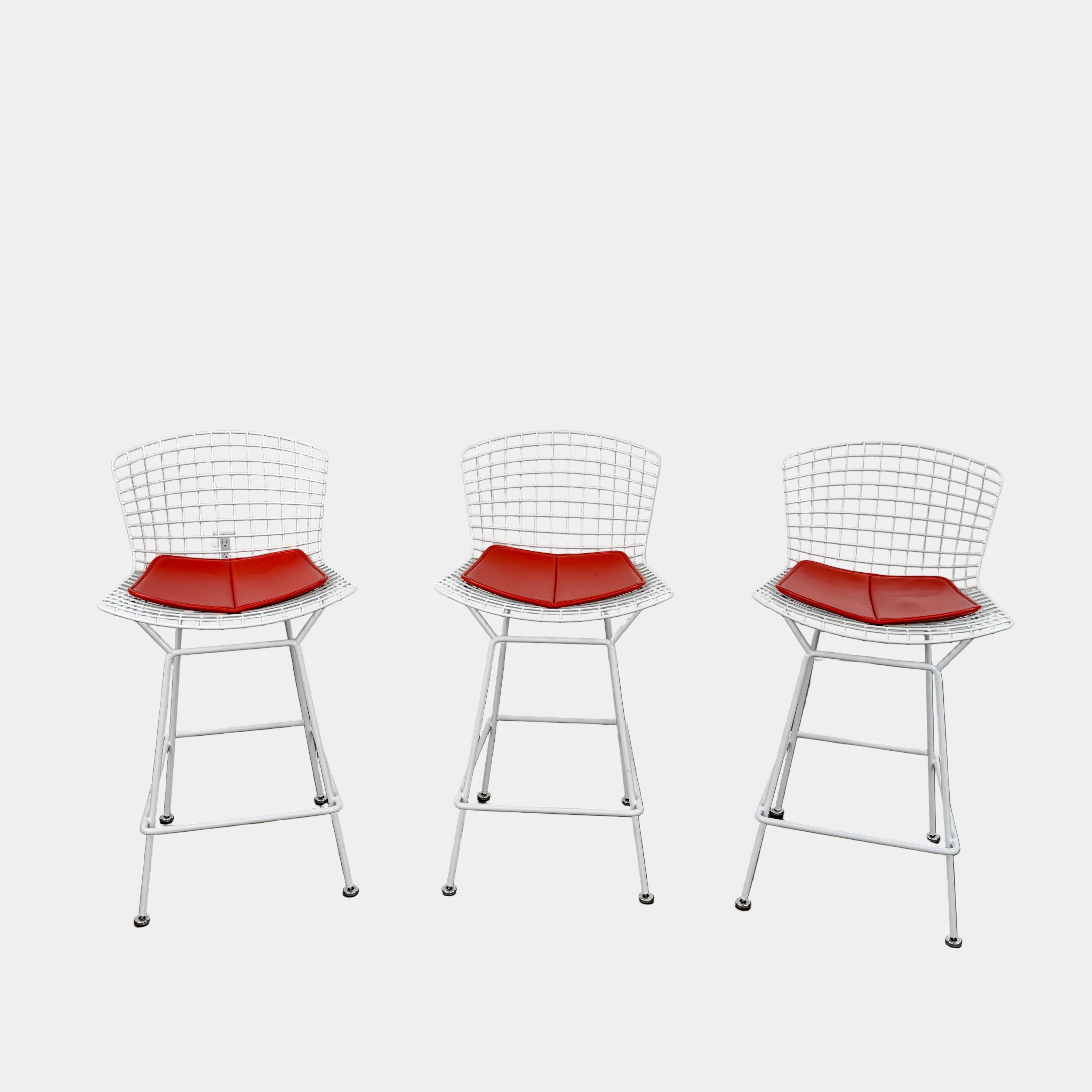 A set of three Bertoia Indoor/Outdoor Counter Stools by Vintage Modern, featuring white welded steel frames with wire backs and bold red seat cushions, lined up in a row against a simple white background—ideal for adding modern style to any indoor or outdoor space.