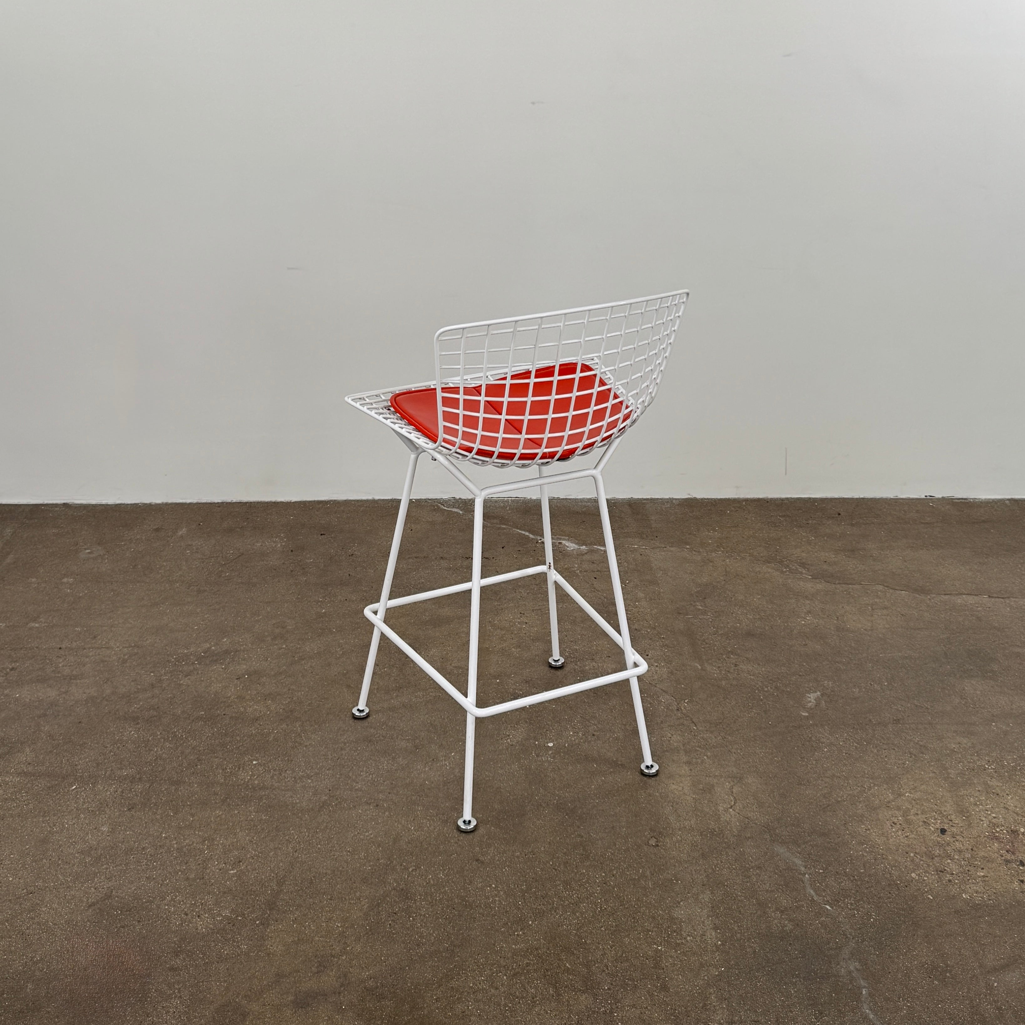 A set of three Bertoia Indoor/Outdoor Counter Stools by Vintage Modern, featuring white welded steel frames with wire backs and bold red seat cushions, lined up in a row against a simple white background—ideal for adding modern style to any indoor or outdoor space.