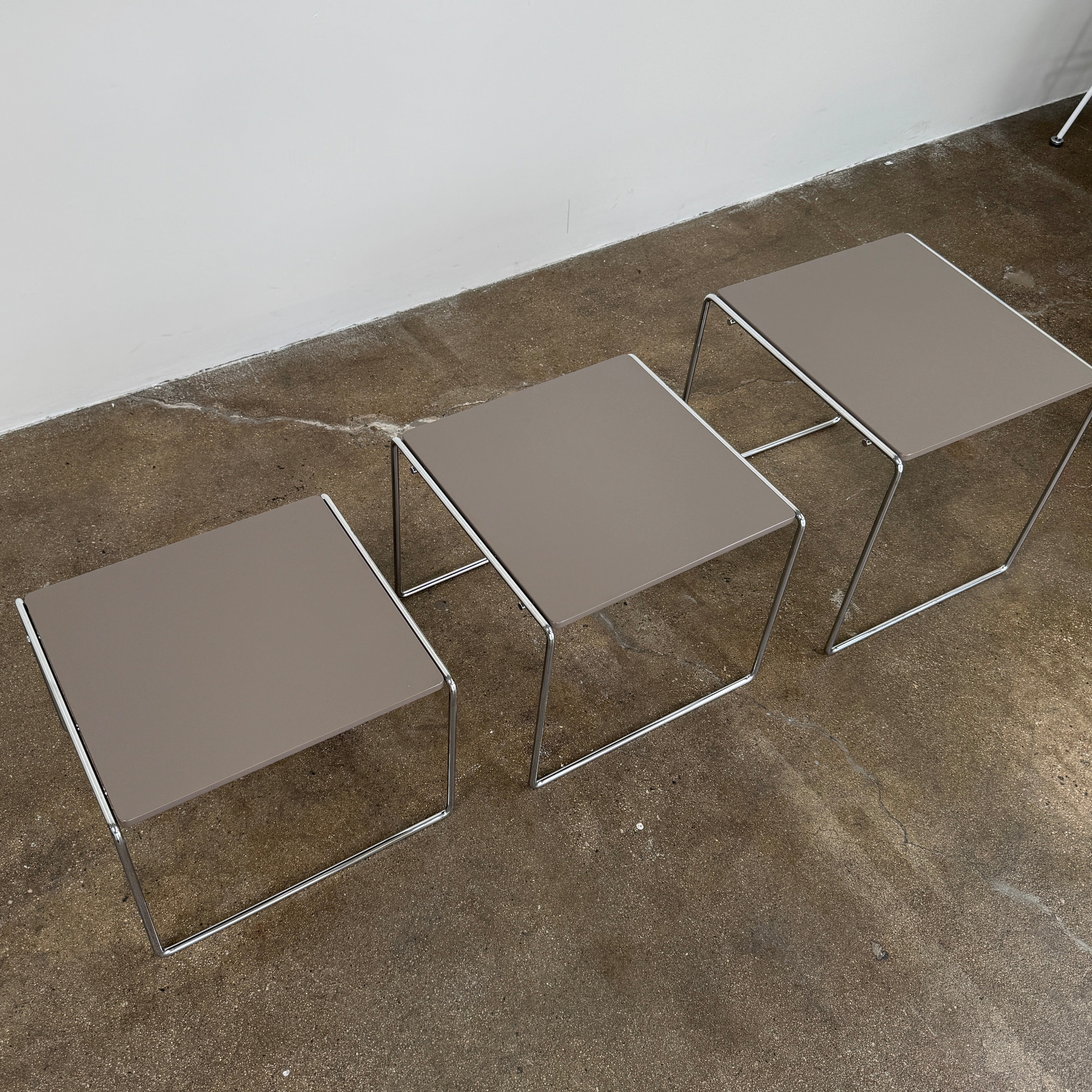 The Cassina Marcel T Set of 3 Nesting Tables by Cassina, featuring taupe tops and sleek metal frames, are displayed in descending order on a concrete floor against a plain white wall, perfectly capturing the essence of modern minimalist design.