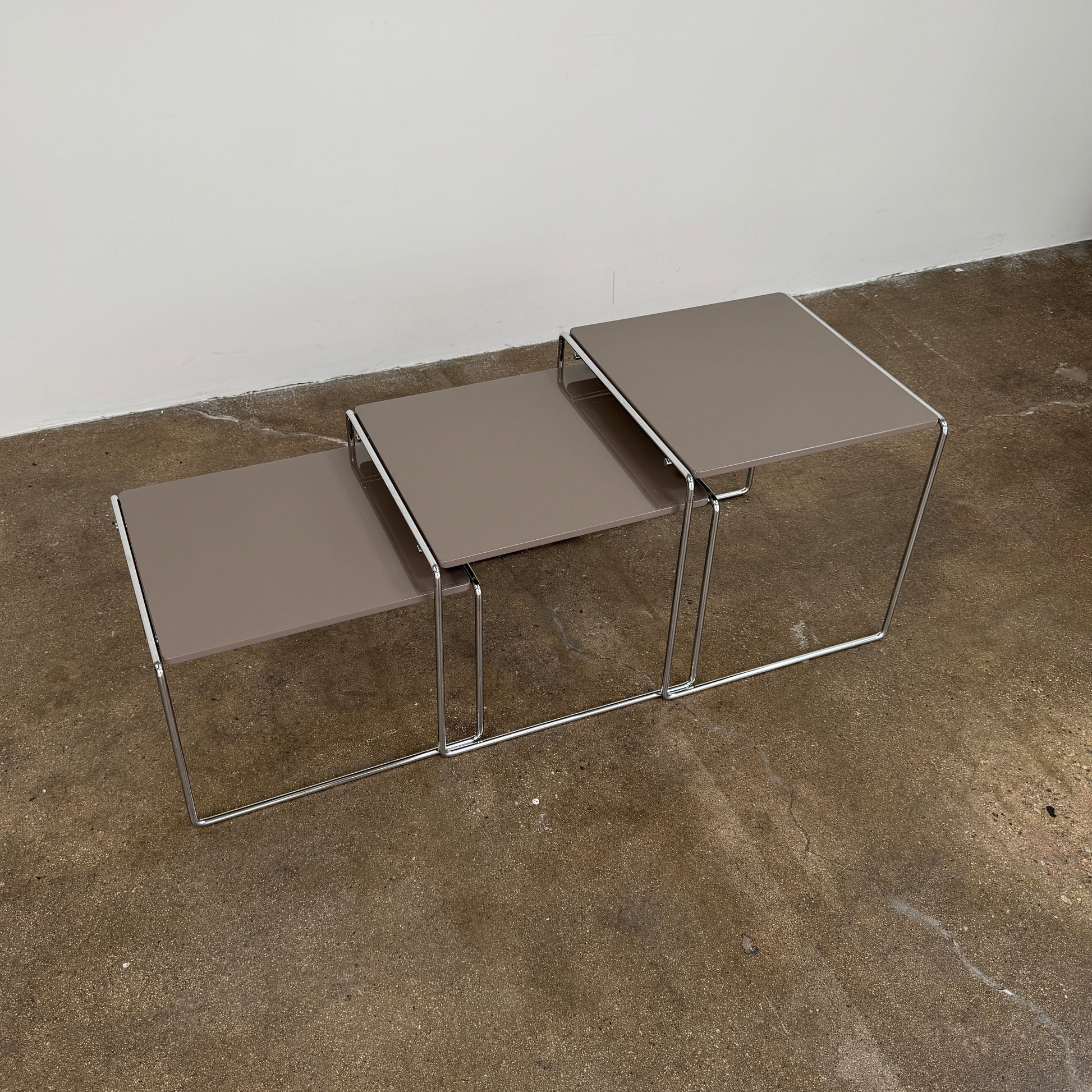 The Cassina Marcel T Set of 3 Nesting Tables by Cassina, featuring taupe tops and sleek metal frames, are displayed in descending order on a concrete floor against a plain white wall, perfectly capturing the essence of modern minimalist design.