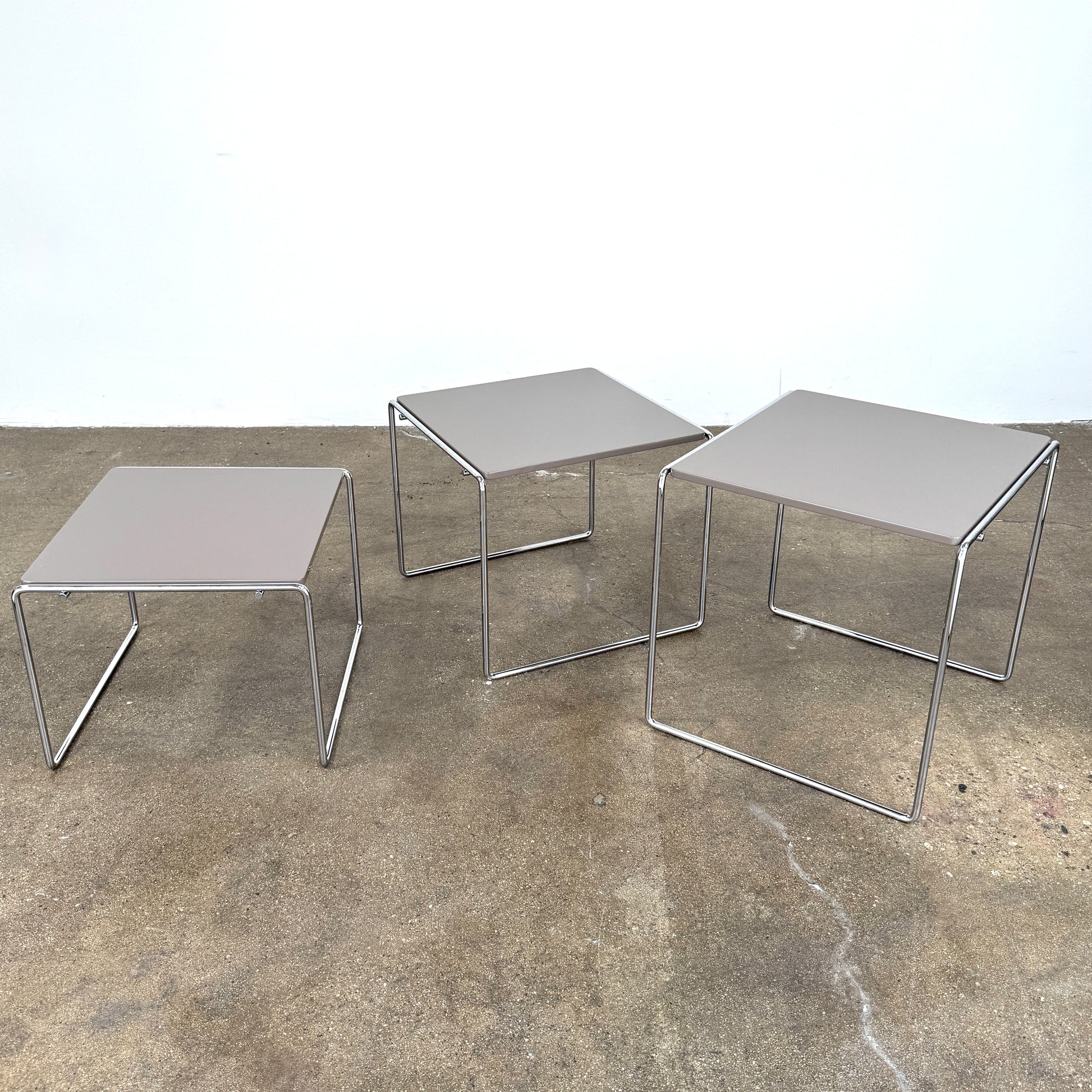 The Cassina Marcel T Set of 3 Nesting Tables by Cassina, featuring taupe tops and sleek metal frames, are displayed in descending order on a concrete floor against a plain white wall, perfectly capturing the essence of modern minimalist design.