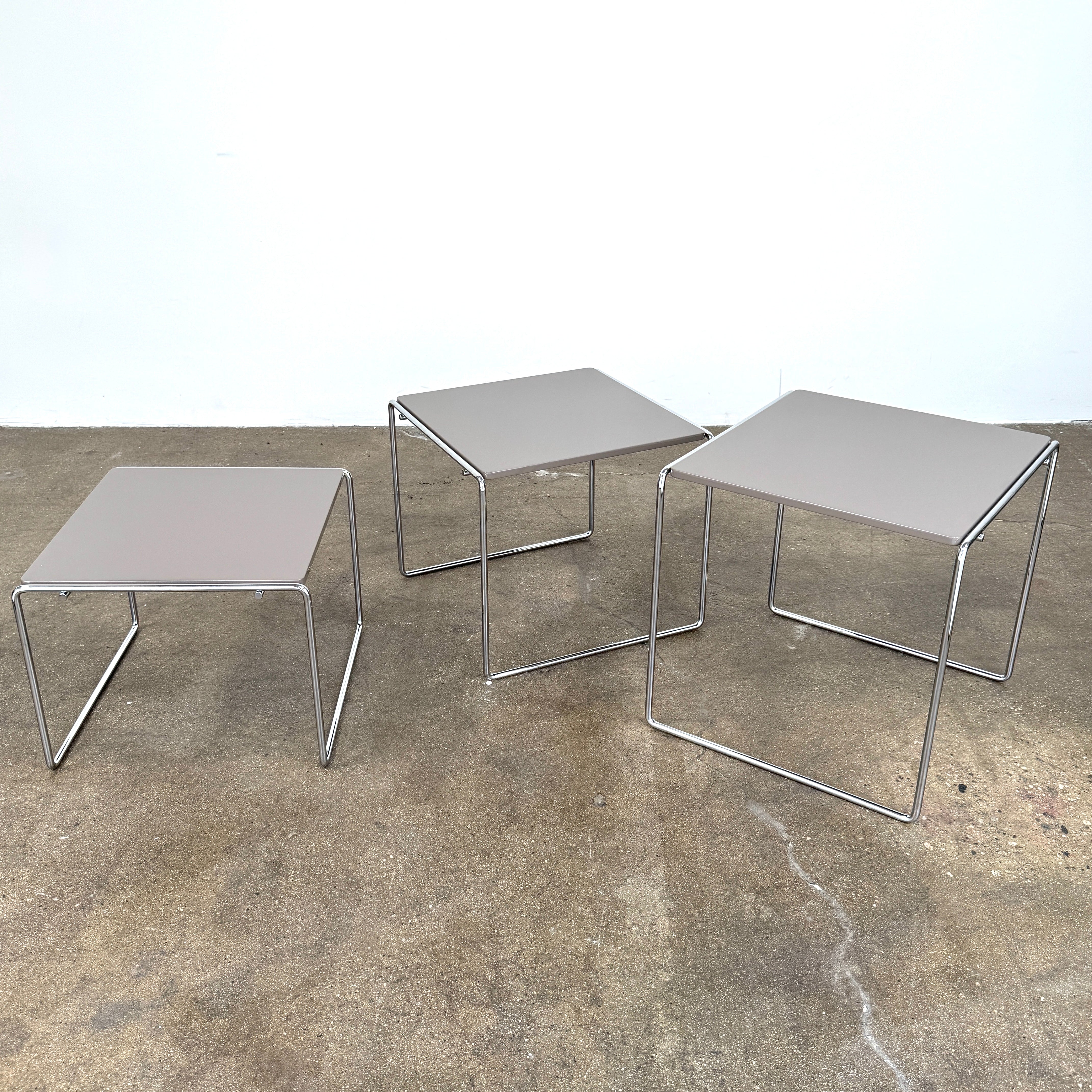 The Cassina Marcel T Set of 3 Nesting Tables by Cassina, featuring taupe tops and sleek metal frames, are displayed in descending order on a concrete floor against a plain white wall, perfectly capturing the essence of modern minimalist design.