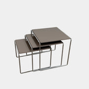 The Cassina Marcel T Set of 3 Nesting Tables by Cassina, featuring taupe tops and sleek metal frames, are displayed in descending order on a concrete floor against a plain white wall, perfectly capturing the essence of modern minimalist design.