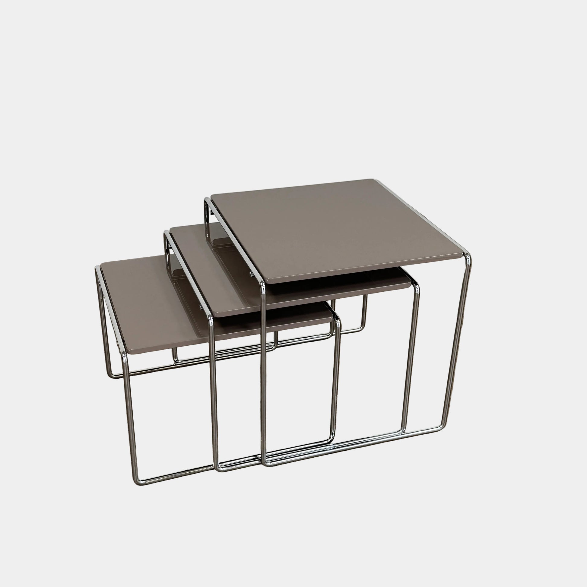The Cassina Marcel T Set of 3 Nesting Tables by Cassina, featuring taupe tops and sleek metal frames, are displayed in descending order on a concrete floor against a plain white wall, perfectly capturing the essence of modern minimalist design.