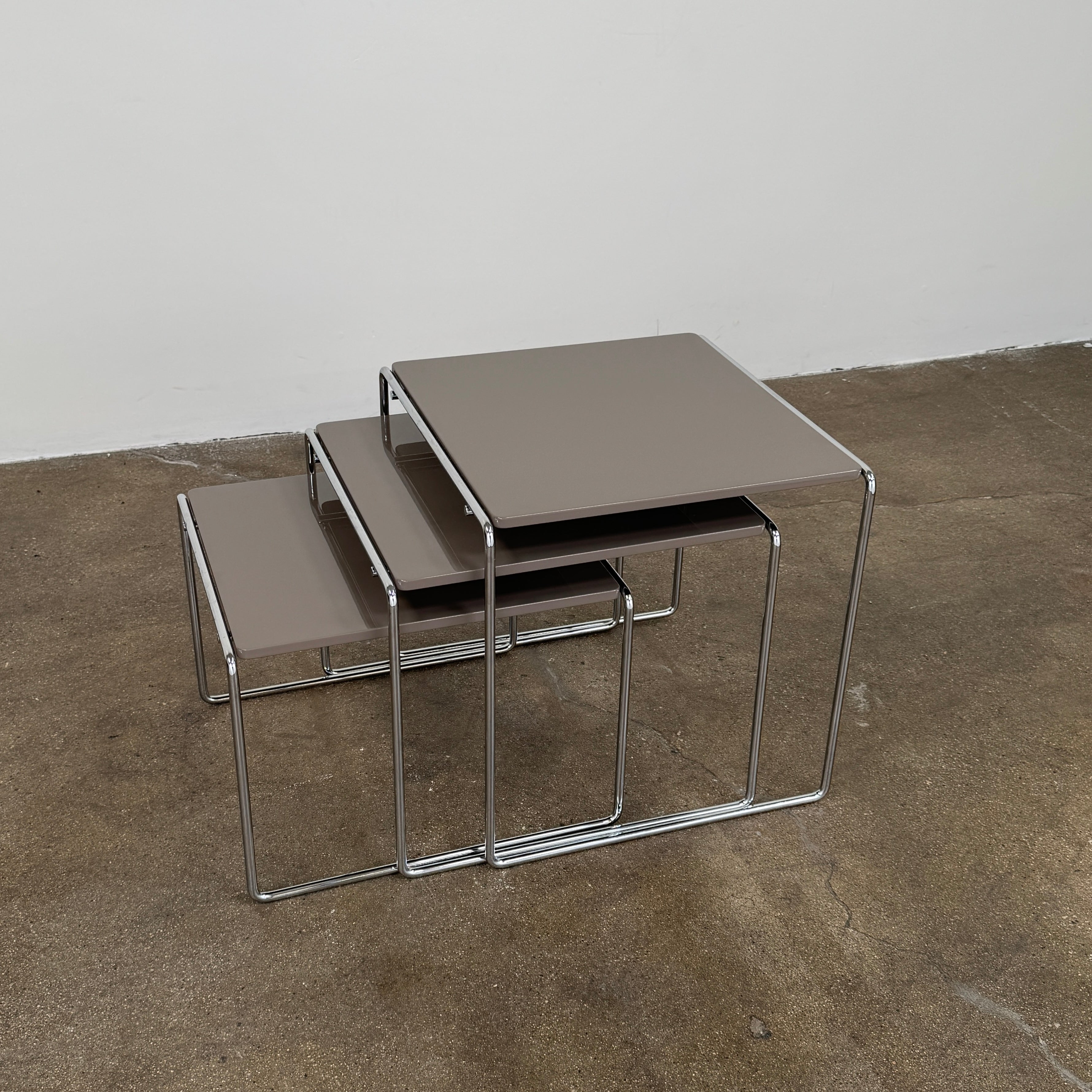 The Cassina Marcel T Set of 3 nesting tables by Modern Resale, featuring taupe tops and metal frames, is displayed on a polished concrete floor against a plain white wall.