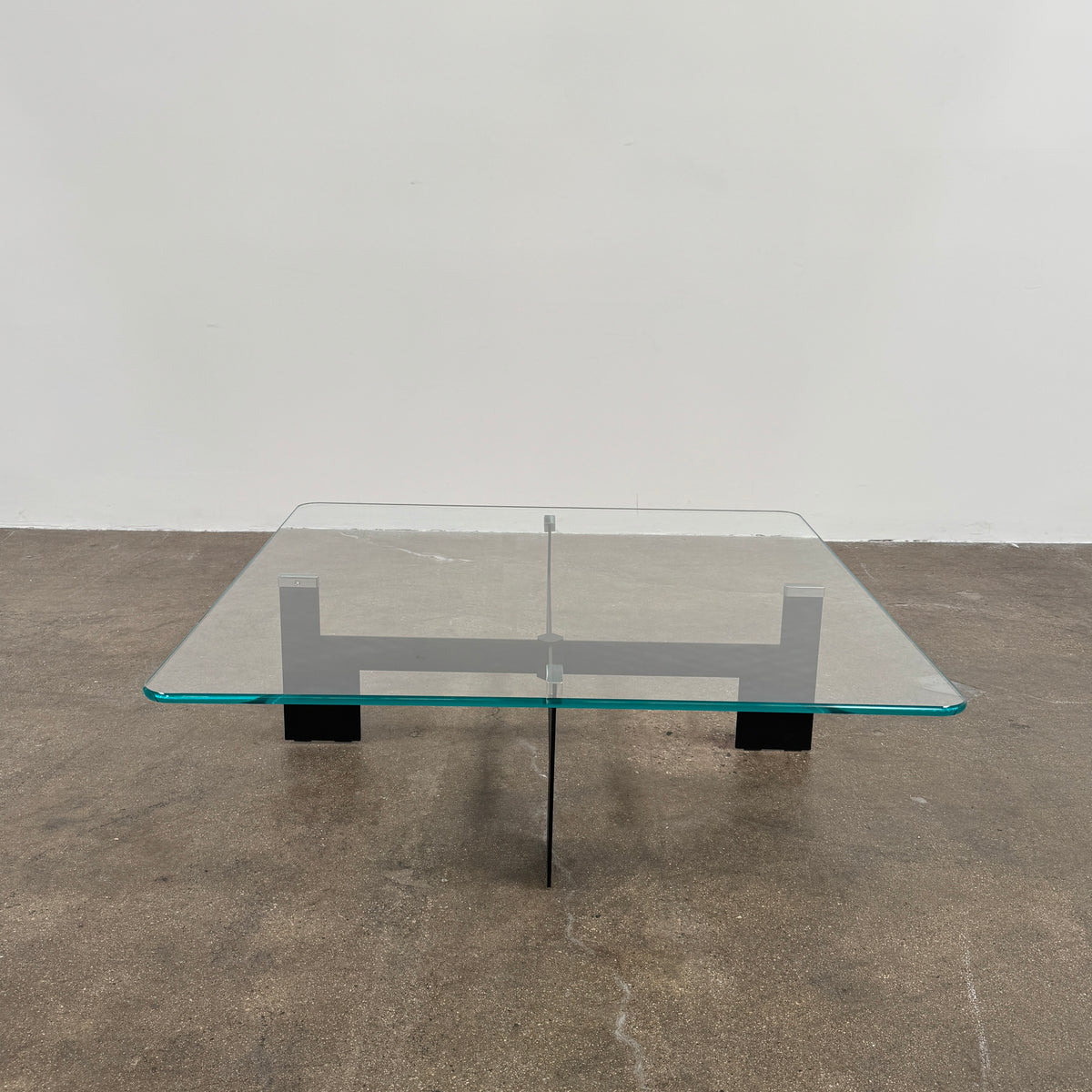 The Minotti Ellis Coffee Table Clear Glass by Minotti features a square clear glass top and a sculptural black metal base, elegantly positioned on a brown concrete floor against a plain white wall.