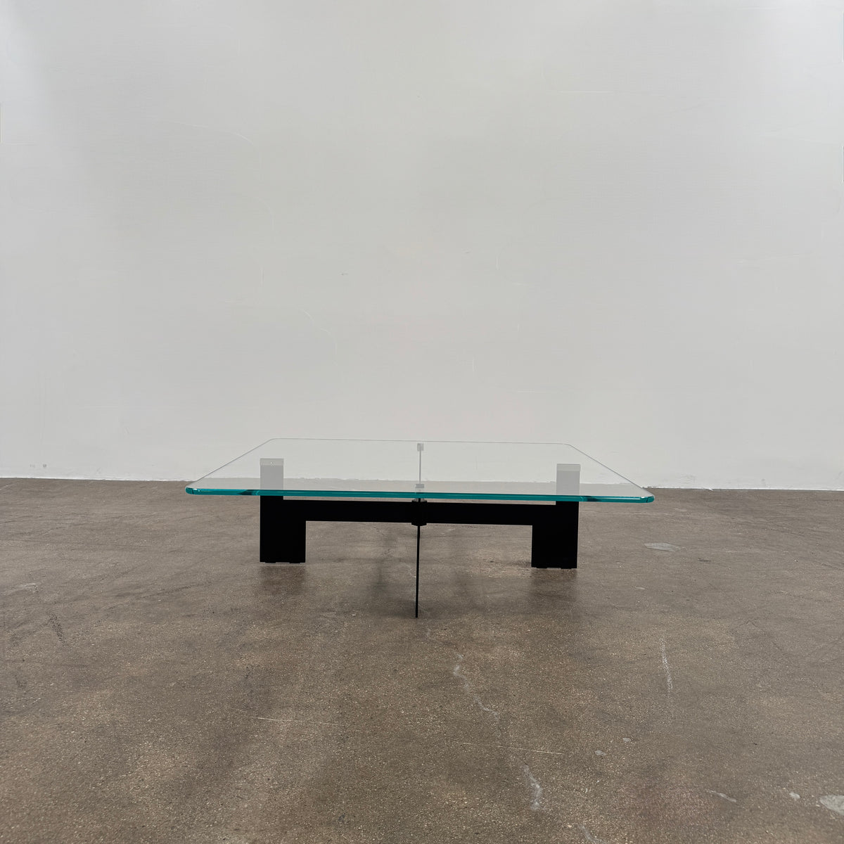 The Minotti Ellis Coffee Table Clear Glass by Minotti features a square clear glass top and a sculptural black metal base, elegantly positioned on a brown concrete floor against a plain white wall.