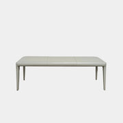 A minimalist, light gray Frag Vella bench by Frag, featuring a padded seat and four legs and handmade in Italy, is positioned against a plain white wall on a brown concrete floor.
