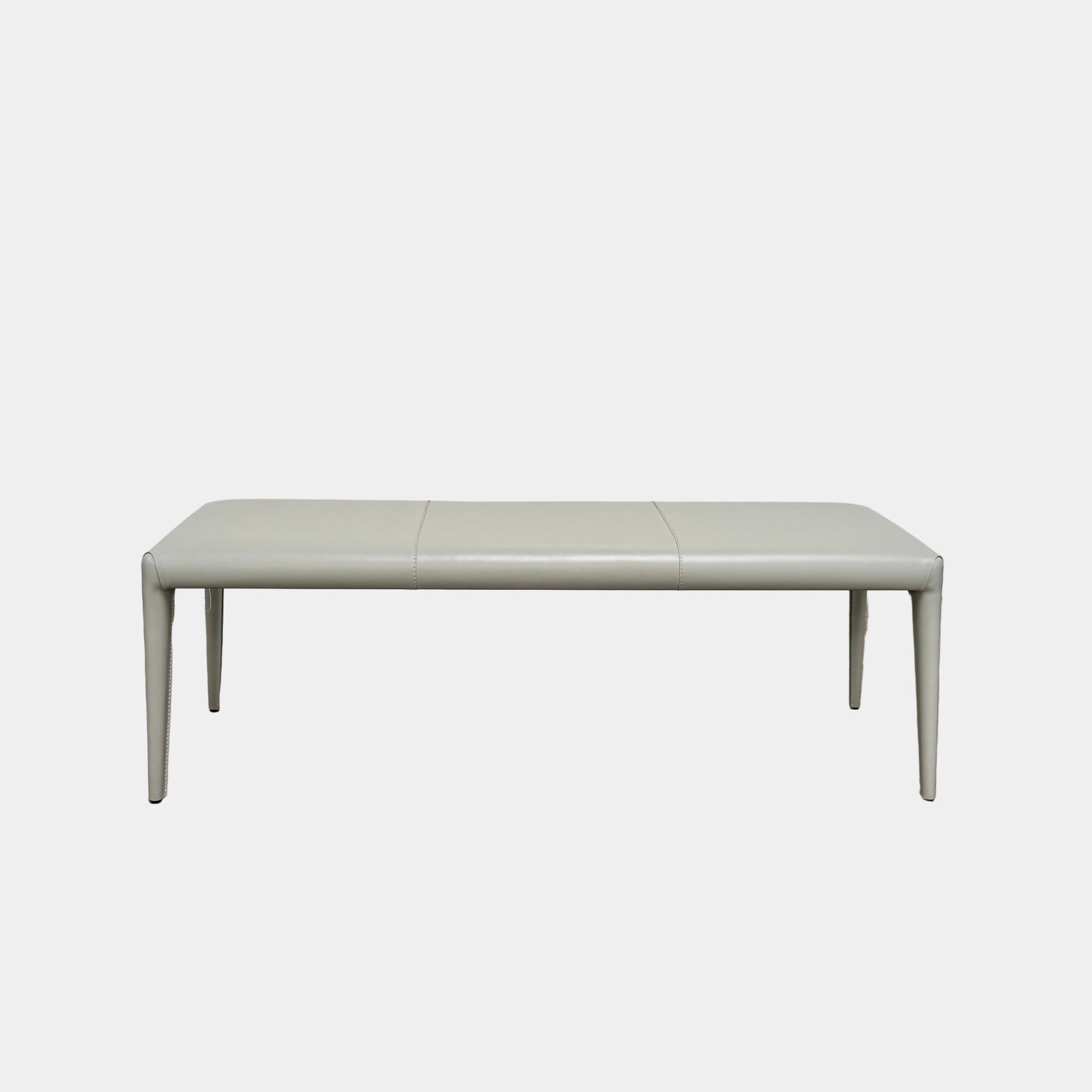 A minimalist, light gray Frag Vella bench by Frag, featuring a padded seat and four legs and handmade in Italy, is positioned against a plain white wall on a brown concrete floor.