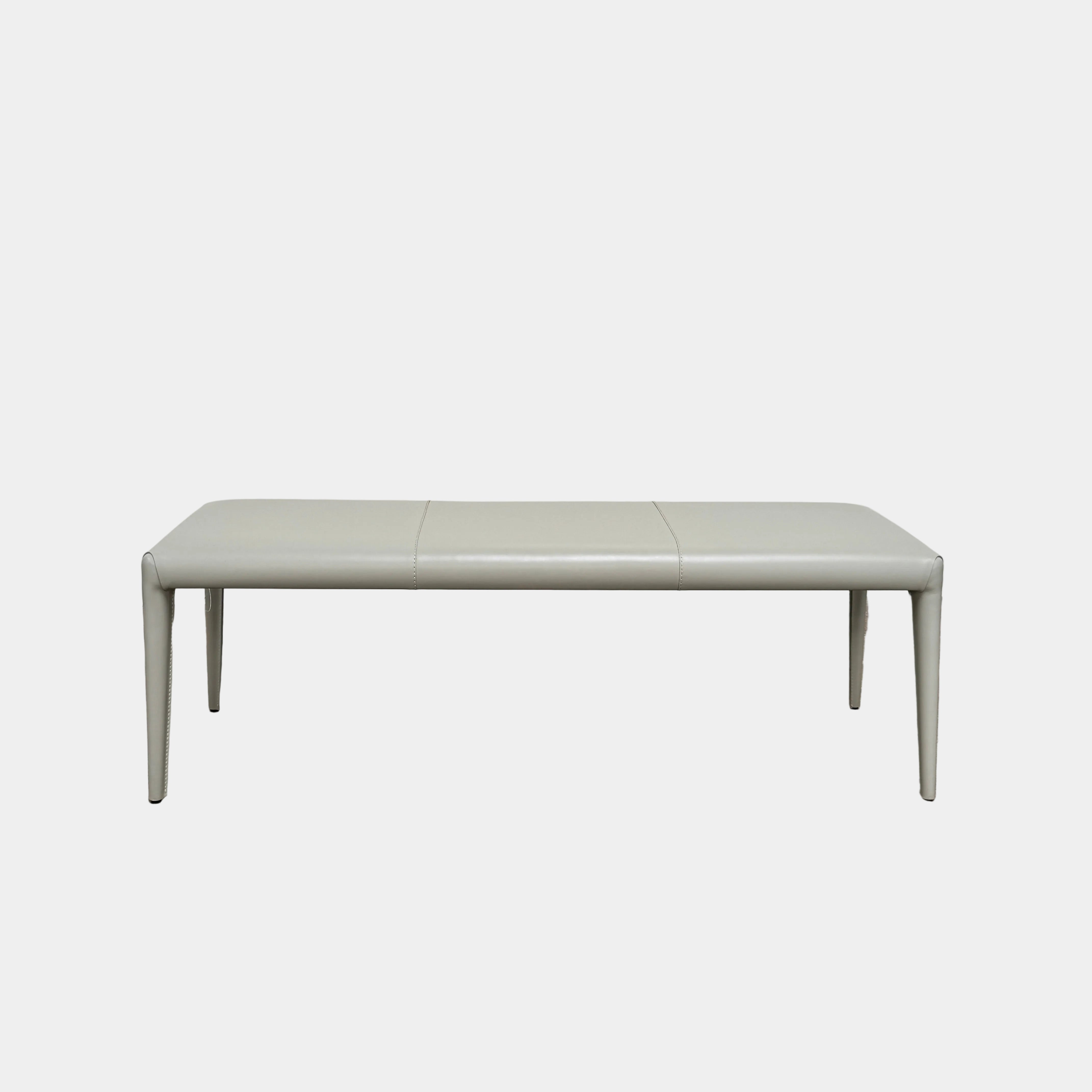 A minimalist, light gray Frag Vella bench by Frag, featuring a padded seat and four legs and handmade in Italy, is positioned against a plain white wall on a brown concrete floor.