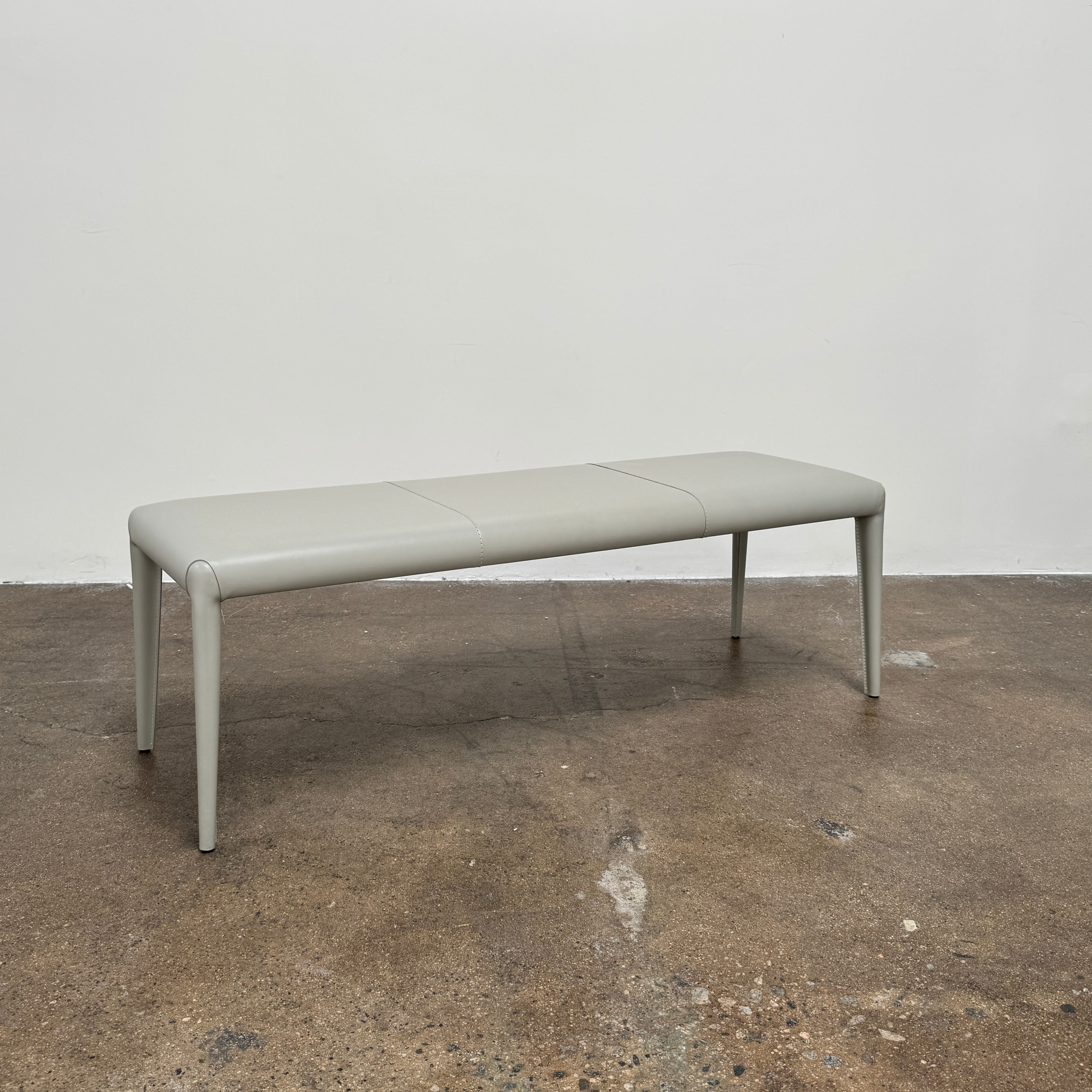 A minimalist, light gray Frag Vella bench by Frag, featuring a padded seat and four legs and handmade in Italy, is positioned against a plain white wall on a brown concrete floor.