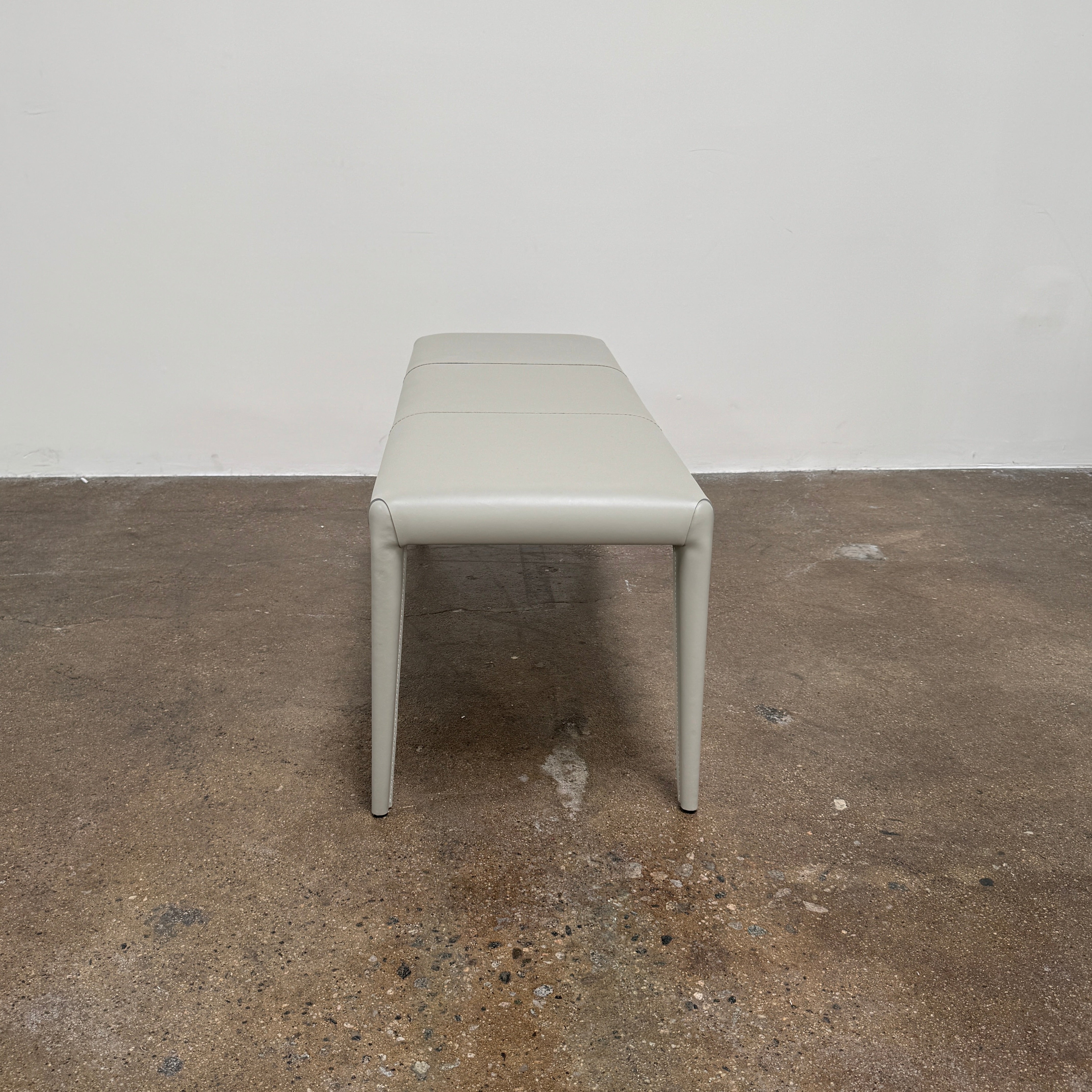 A minimalist, light gray Frag Vella bench by Frag, featuring a padded seat and four legs and handmade in Italy, is positioned against a plain white wall on a brown concrete floor.